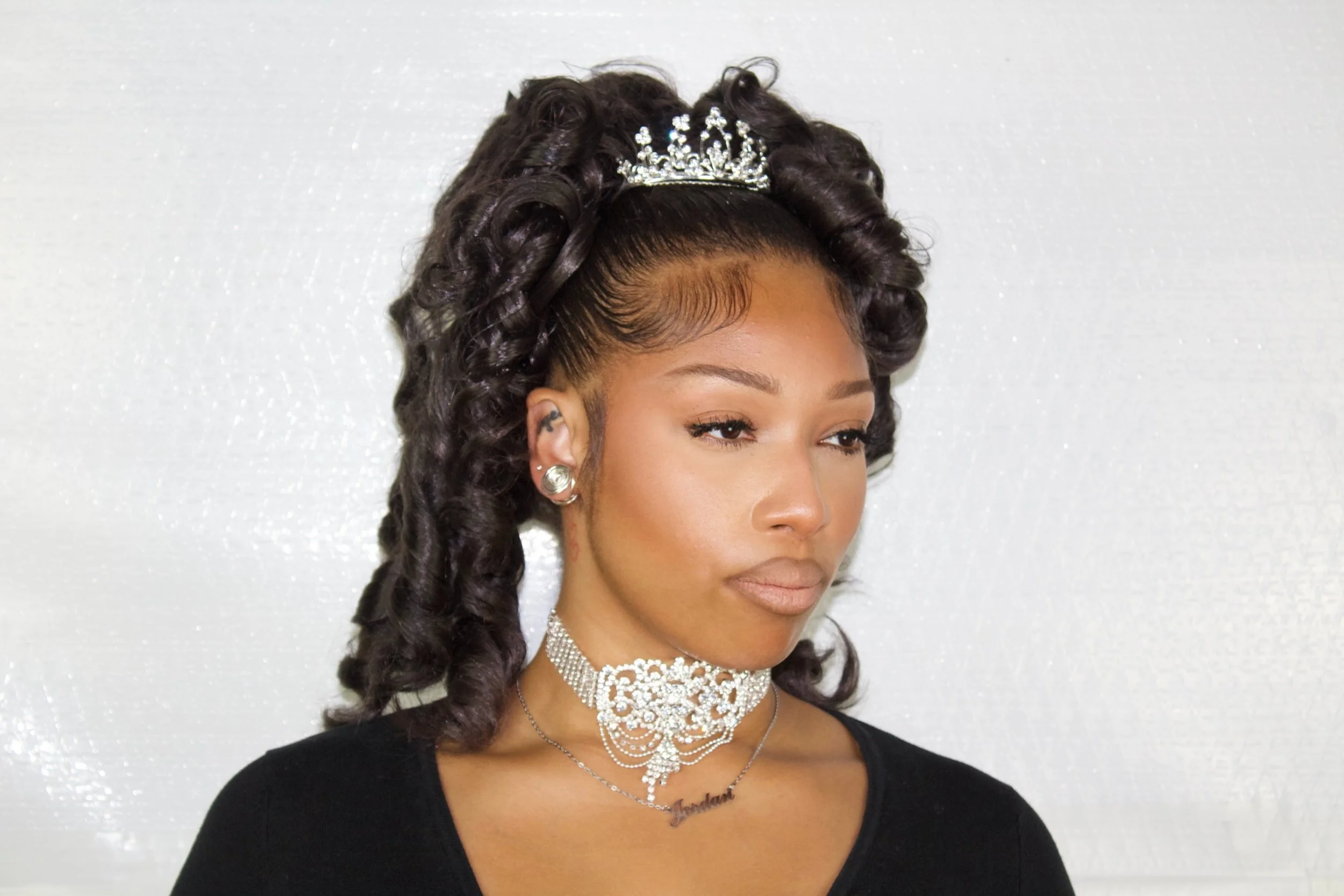 A woman with styled dark curly hair wearing a tiara, large earrings, a diamond necklace, and a black top, standing against a plain light-colored background.
