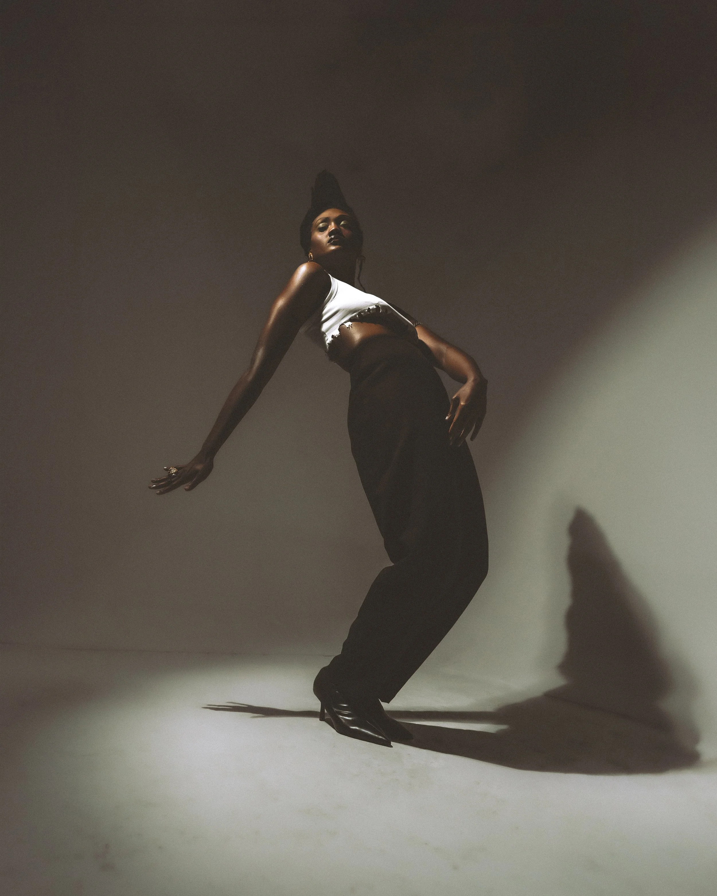 A woman with dark skin poses in a studio with a gradient background, wearing a white cropped top and black pants, with heels and gold earrings, leaning back with one arm extended and the other on her hip.