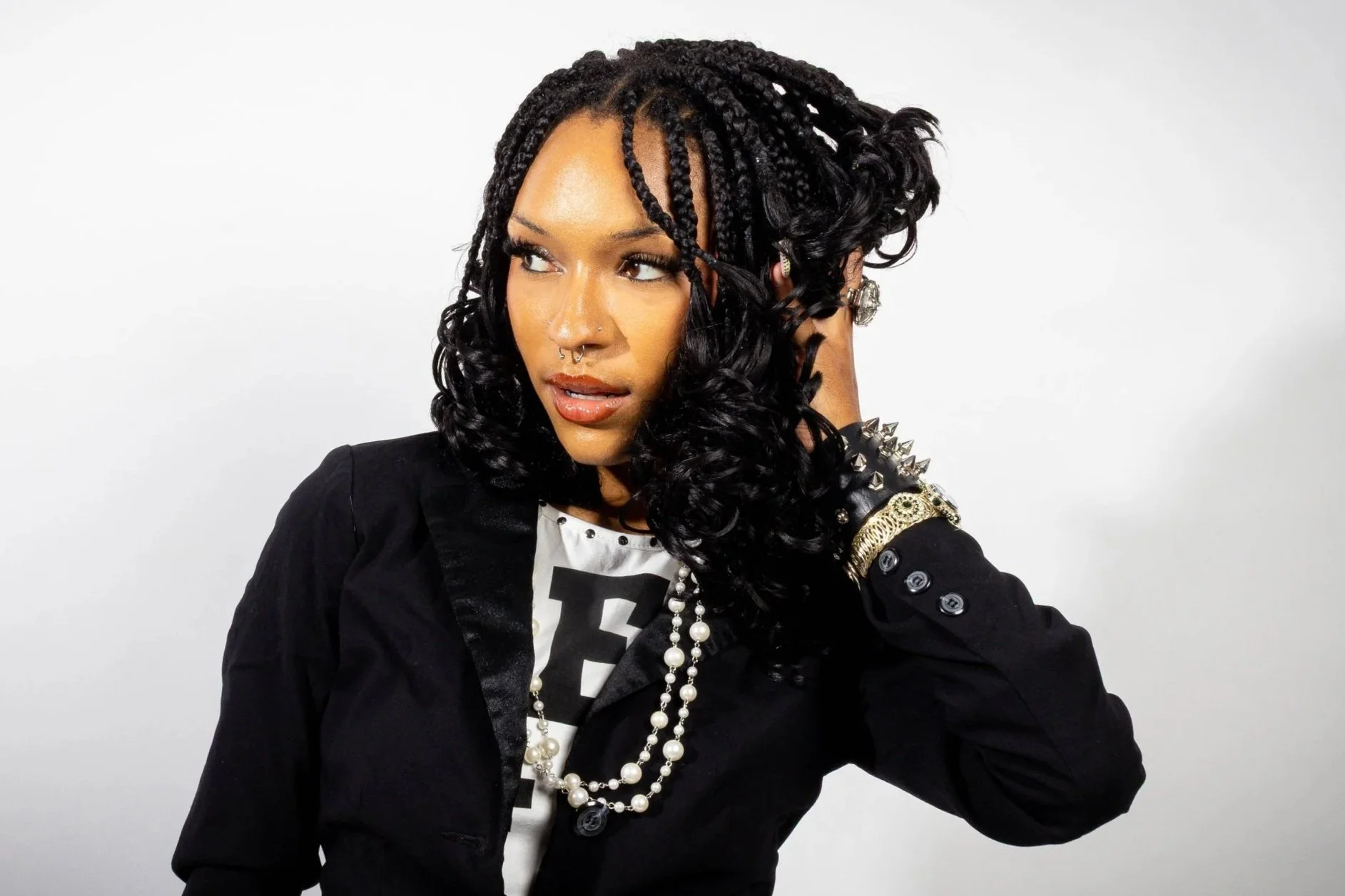 A woman with styled curly hair and braids, wearing a black jacket with spikes on the shoulders, multiple pearl necklaces, and earrings, posing against a plain white background.