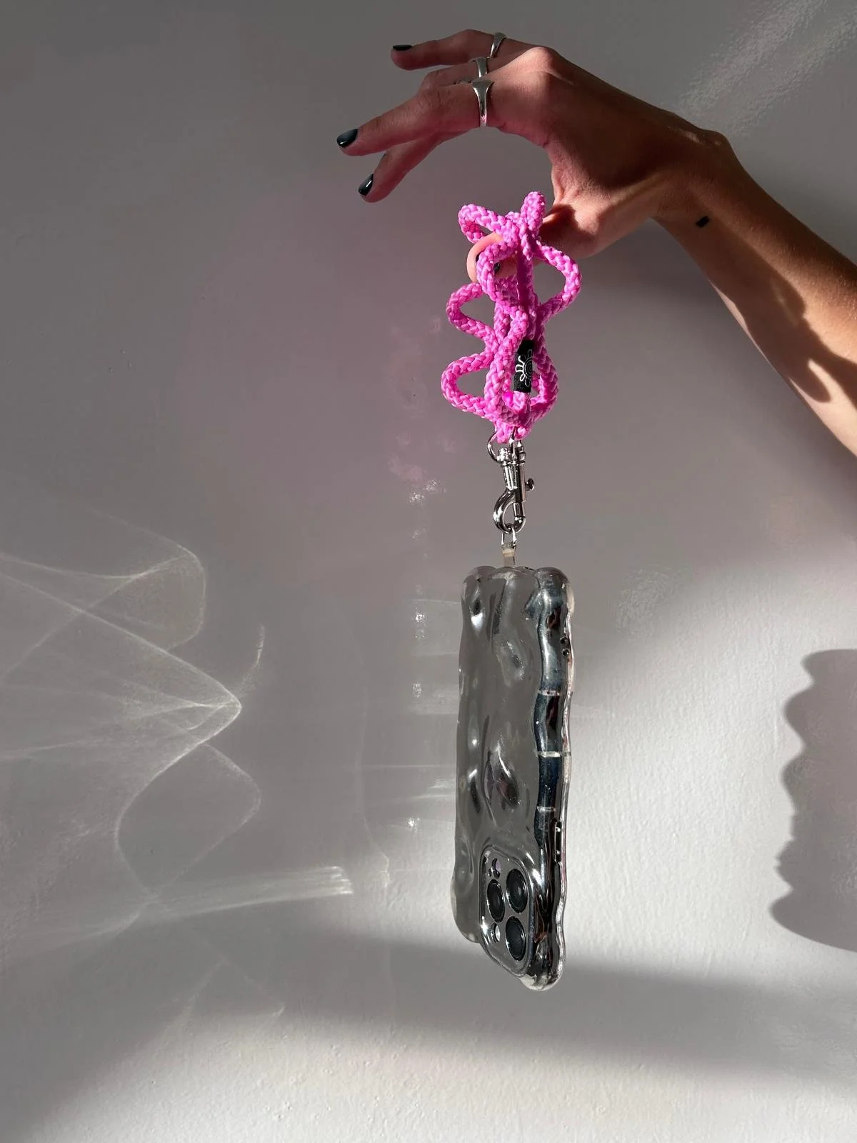 Hand holding a reflective metallic phone case with a pink cord attached, against a plain wall.