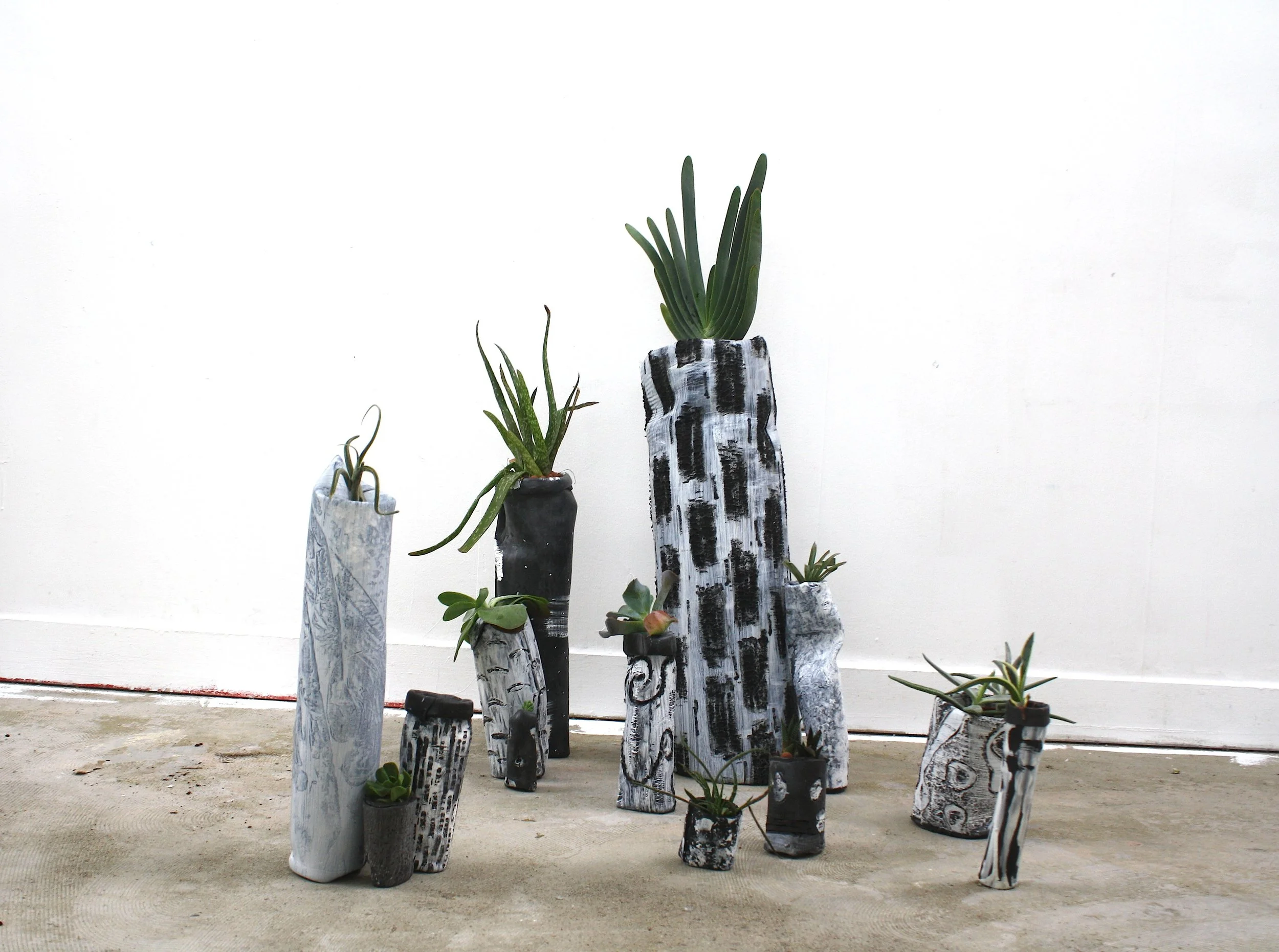 Collection of tall, painted ceramic vases with succulents and plants inside, arranged on a concrete floor against a plain white wall.