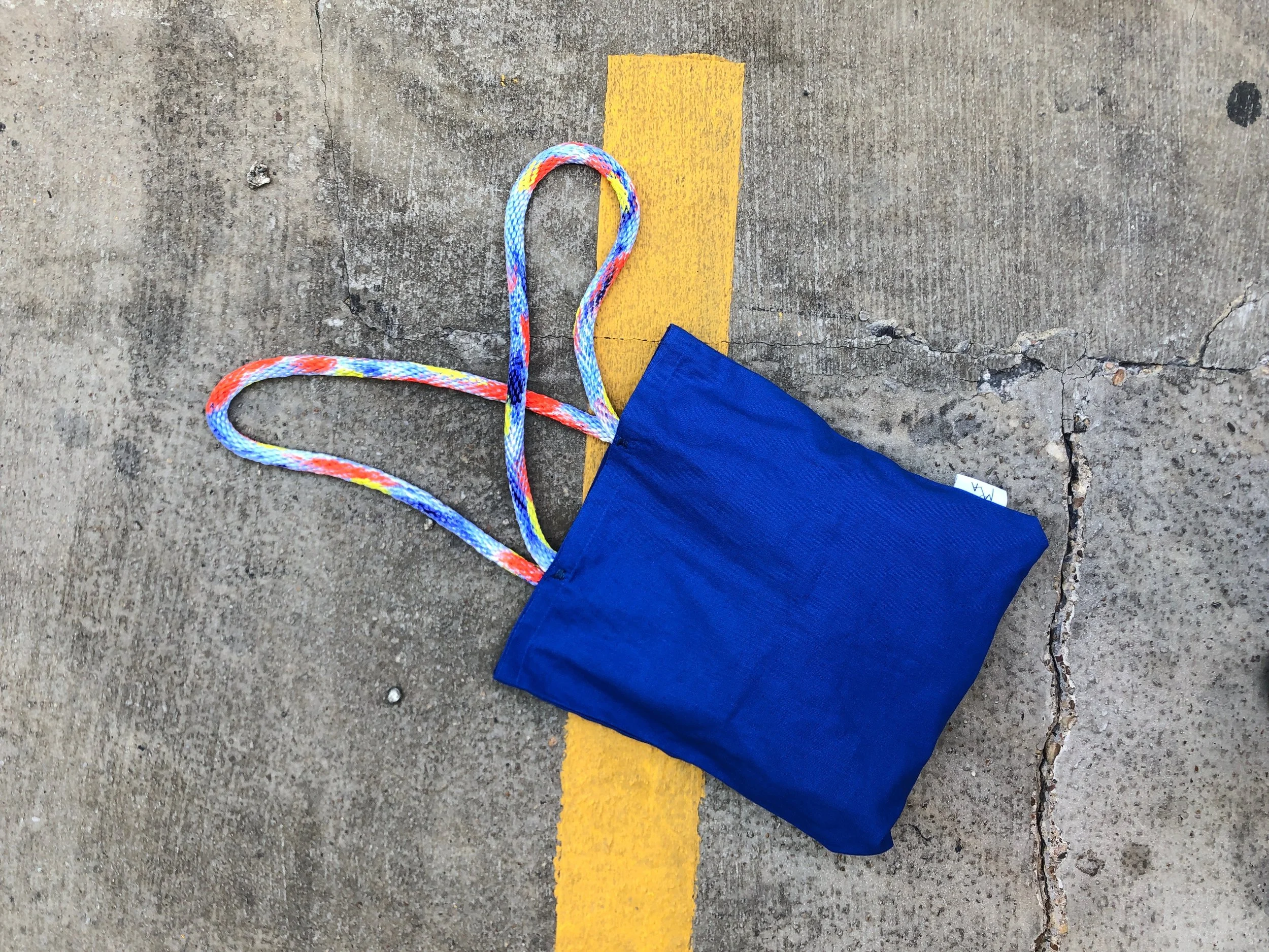 A blue cloth bag with multicolored striped handle resting on a concrete surface with a yellow painted line.