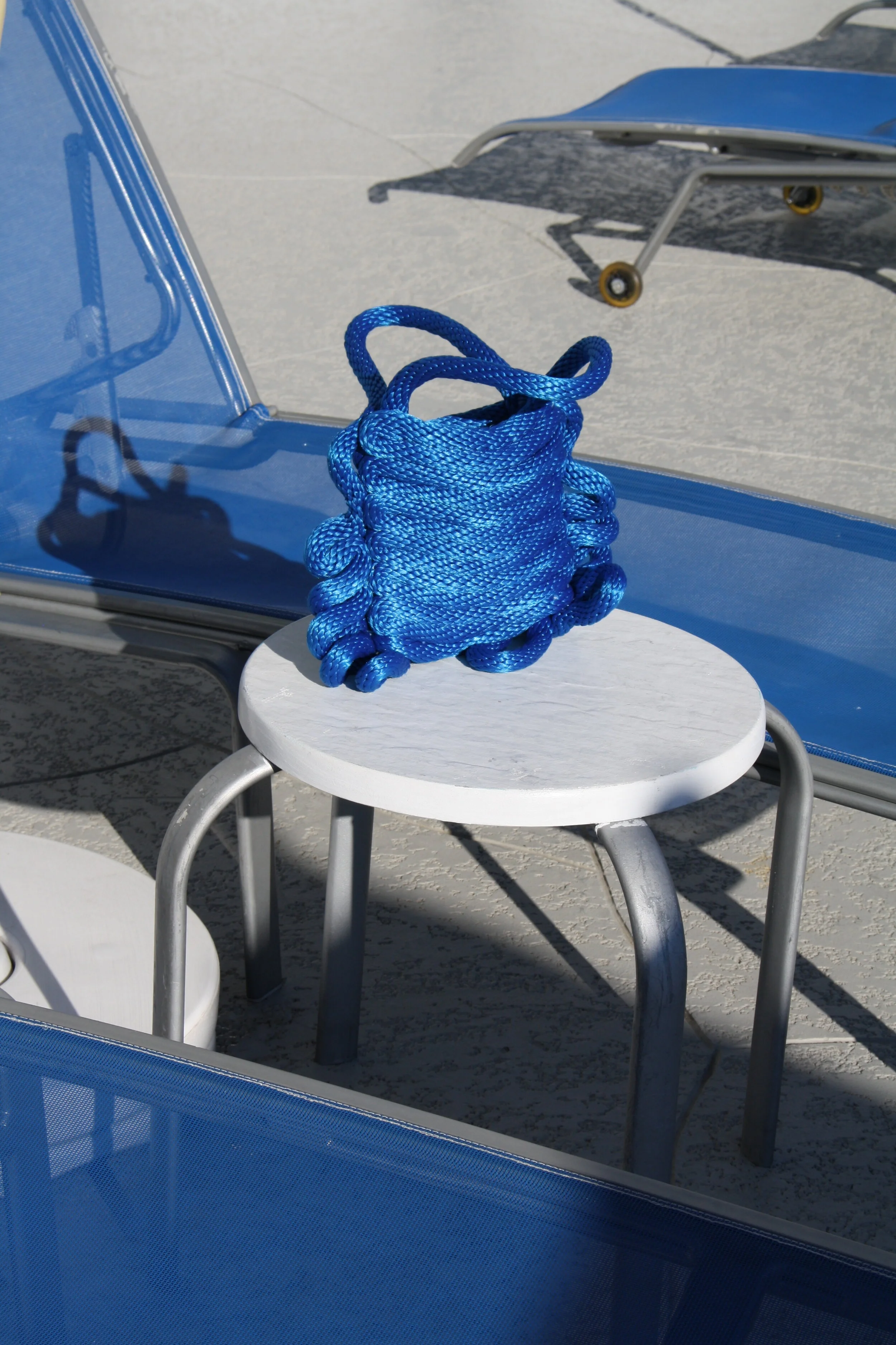 A blue rope sculpture resembling a frog sits on a small, round white table outdoors, with lounge chairs in the background.