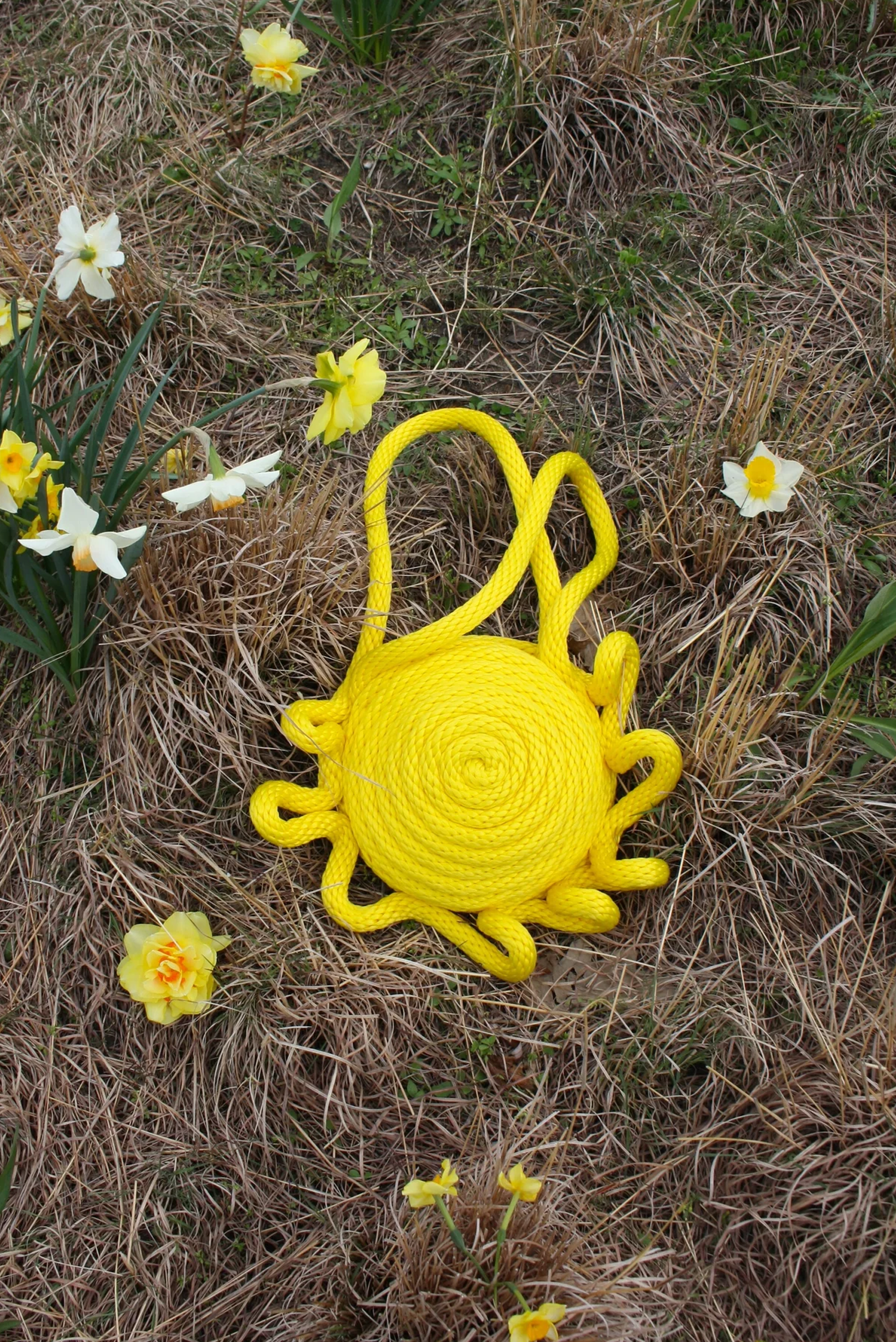 Yellow rope art arranged in the shape of a sun with curled rays, lying on dry grass surrounded by yellow and white flowers.
