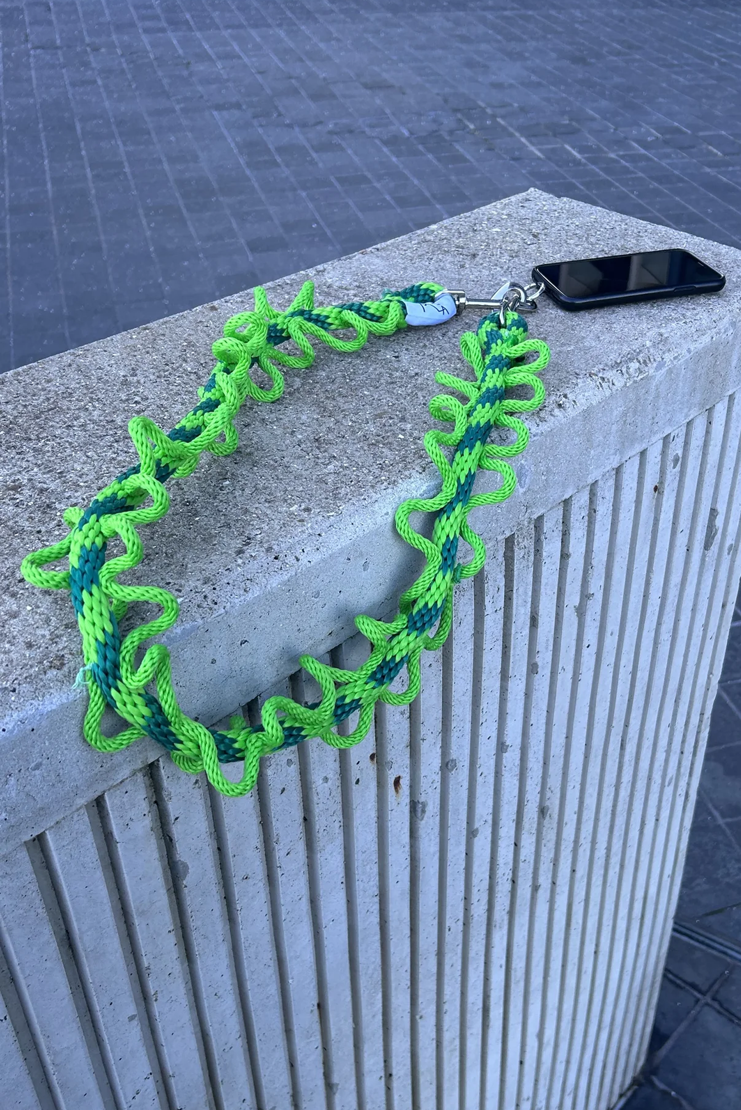 Neon green and blue paracord necklace with a small white tag, attached to a black smartphone, resting on a concrete ledge in an outdoor setting.