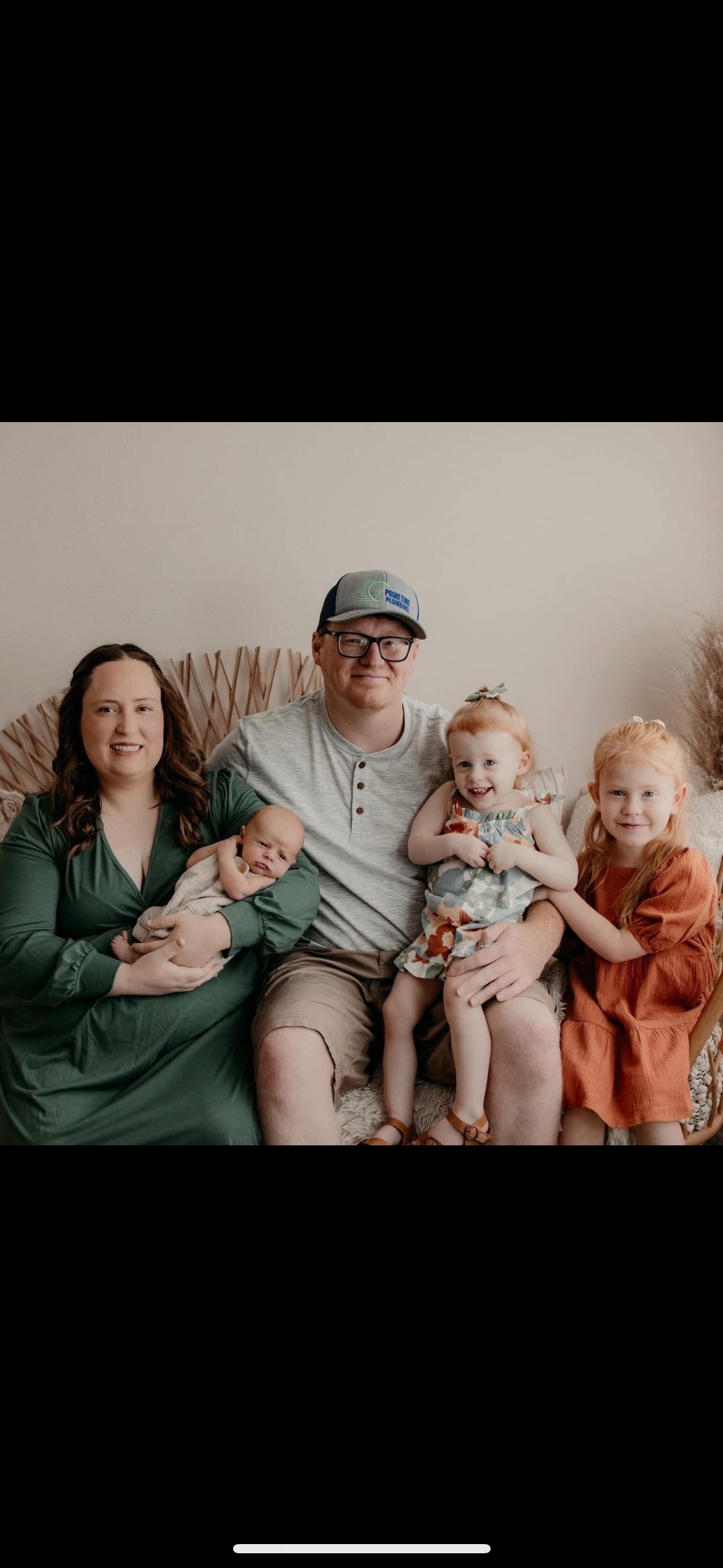 A family of five sitting together on a couch, including a woman holding a baby, a man, and three young girls. The woman has long brown hair and is wearing a green dress. The man is wearing glasses, a gray hat, and a gray shirt. The baby has a light outfit and is being held by the woman. The two younger girls are sitting on the man's lap, smiling, with one wearing a colorful dress and the other in an orange dress.