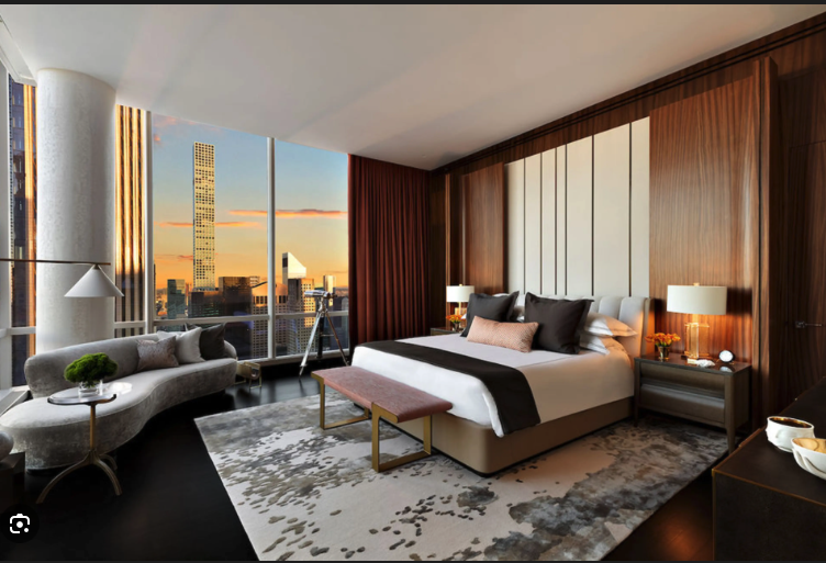 Modern bedroom with large window view of city skyline at sunset, featuring a king-sized bed with white and dark gray bedding, wooden accent wall, bedside tables with lamps, a curved sofa with cushions, a coffee table, and a geometric rug.