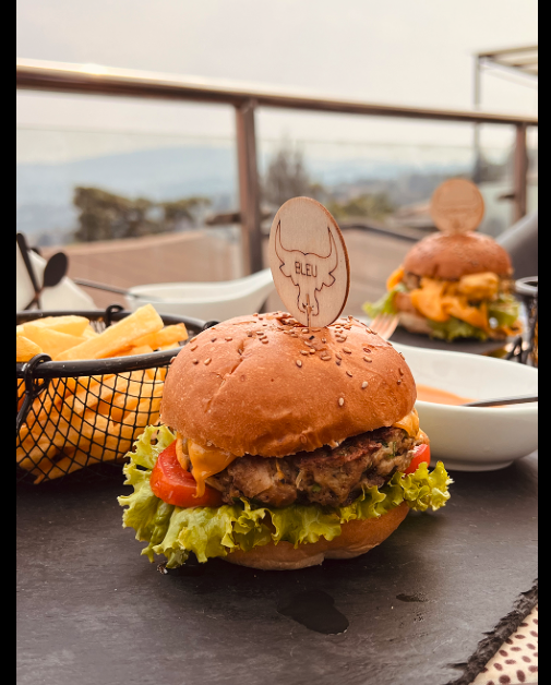 Burger with lettuce, tomato, melted cheese, and a sesame seed bun, served on a black table with a side of fries and another burger in the background.