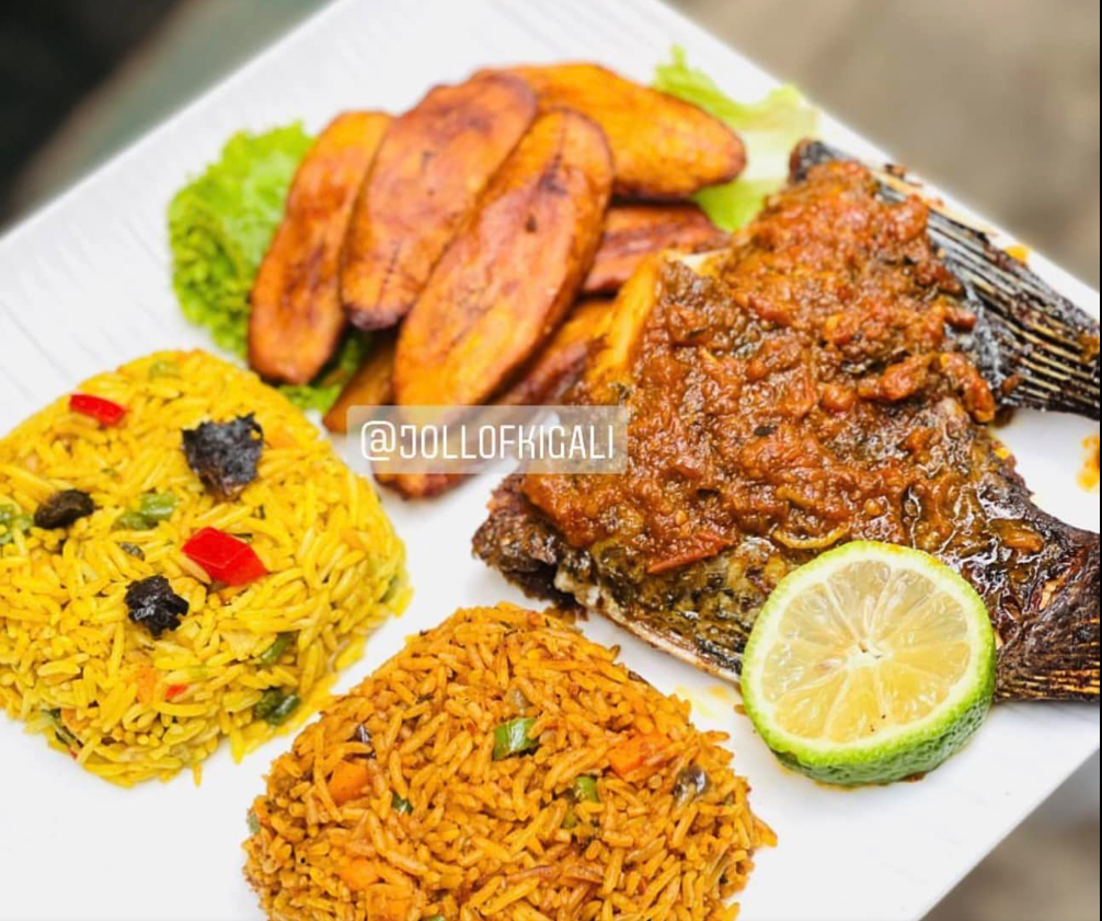 Plate of grilled fish topped with tomato sauce, served with yellow rice, jollof rice, fried plantains, and a lemon wedge.