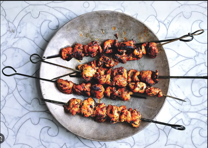 Four skewers of grilled chicken satay with charred edges on a round metal plate.