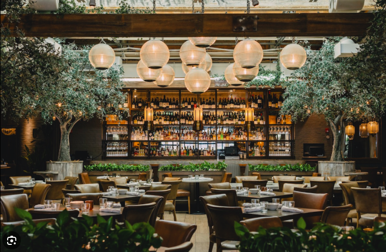 Elegant restaurant interior with a full bar, modern lighting, multiple tables set for dining, and lush greenery.