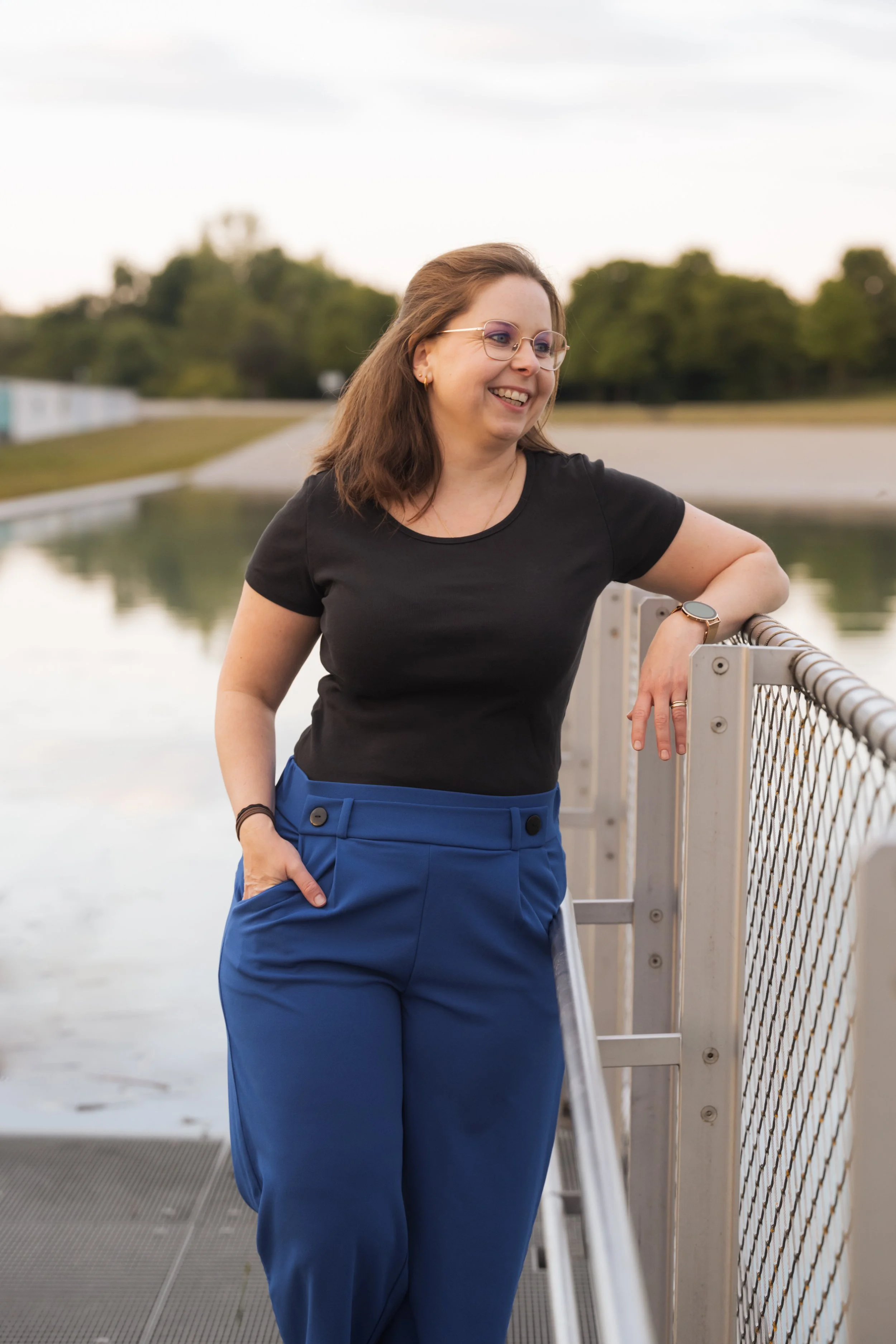 Eine lächelnde Frau mit Brille, schwarzen T-Shirt und blauen Hosen lehnt an einem Metallgeländer an einem See mit grünem Hintergrund.