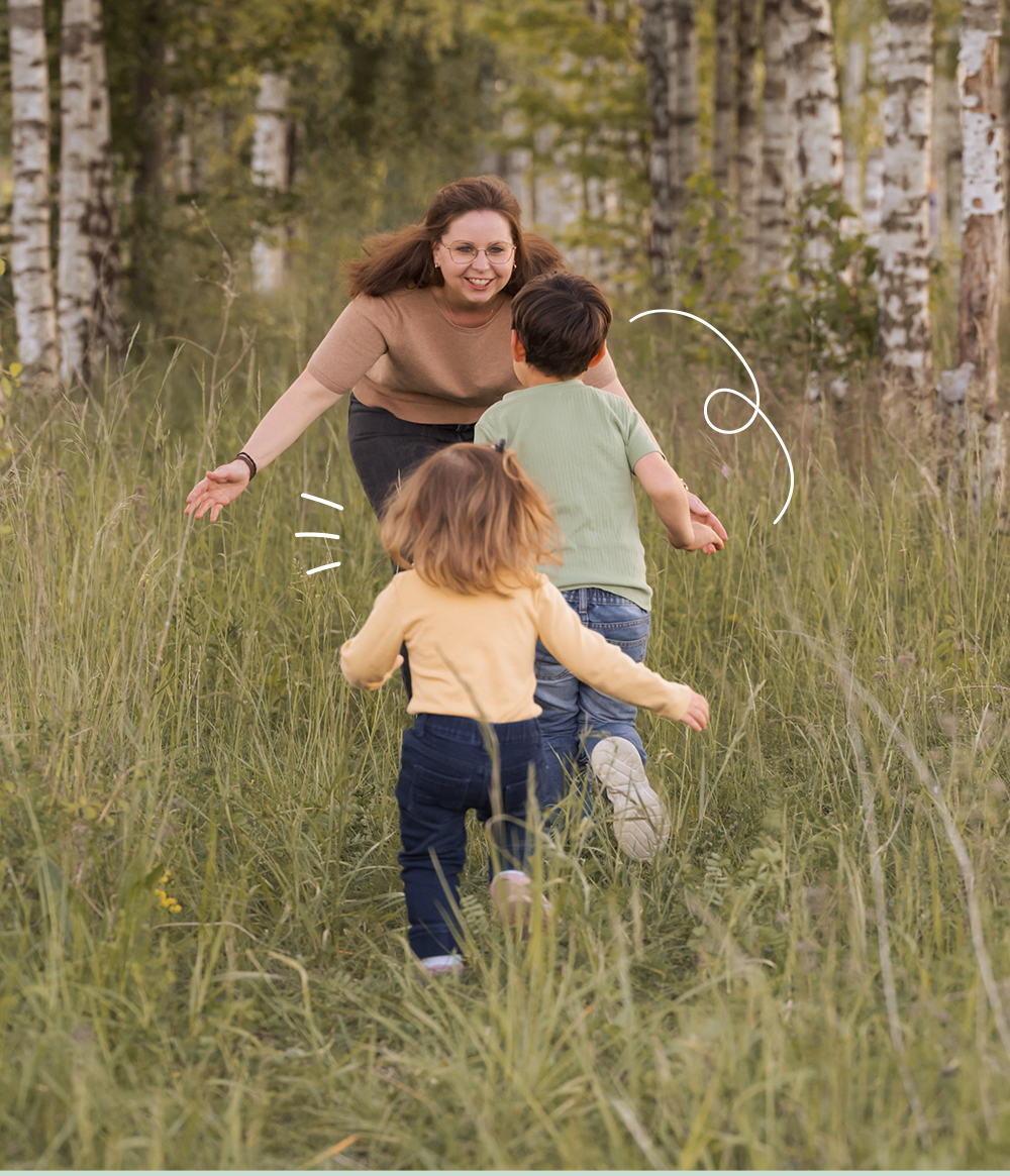 Eine lachende Frau läuft lachend mit zwei Kindern durch einen Wald im Sommer, während sie zusammen spielen.