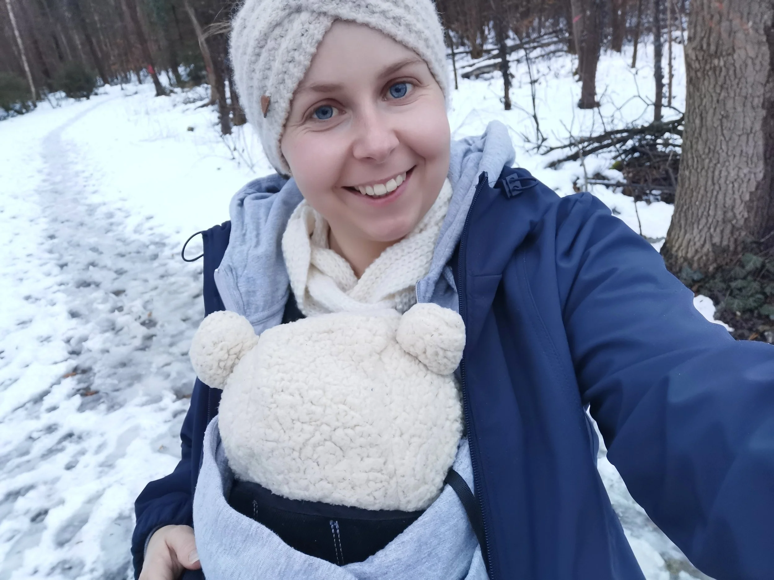 Eine Frau mit blauen Augen, braunem Haar unter einem gestrickten Wollhut, die im Schnee in einem Wald steht, trägt eine dunkelblaue Jacke, einen weißen Schal und eine hellbraune Mütze mit Ohren. Sie hält ein schlafendes Baby, das eine hellbraune, kuschelige Mütze mit Ohren trägt.