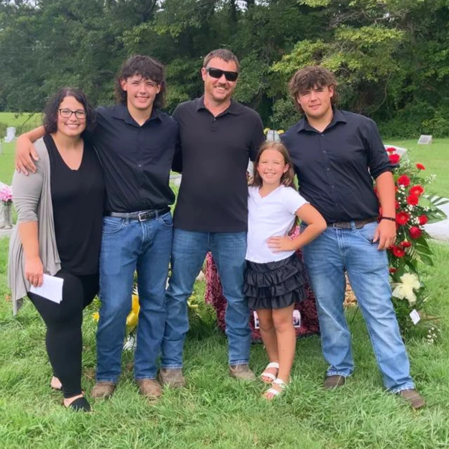 A family of five standing outdoors on grass, smiling, with trees in the background. They are dressed in casual clothes, with three males in black shirts and two females, one in a black top and gray cardigan, and a young girl in a white top and black skirt.