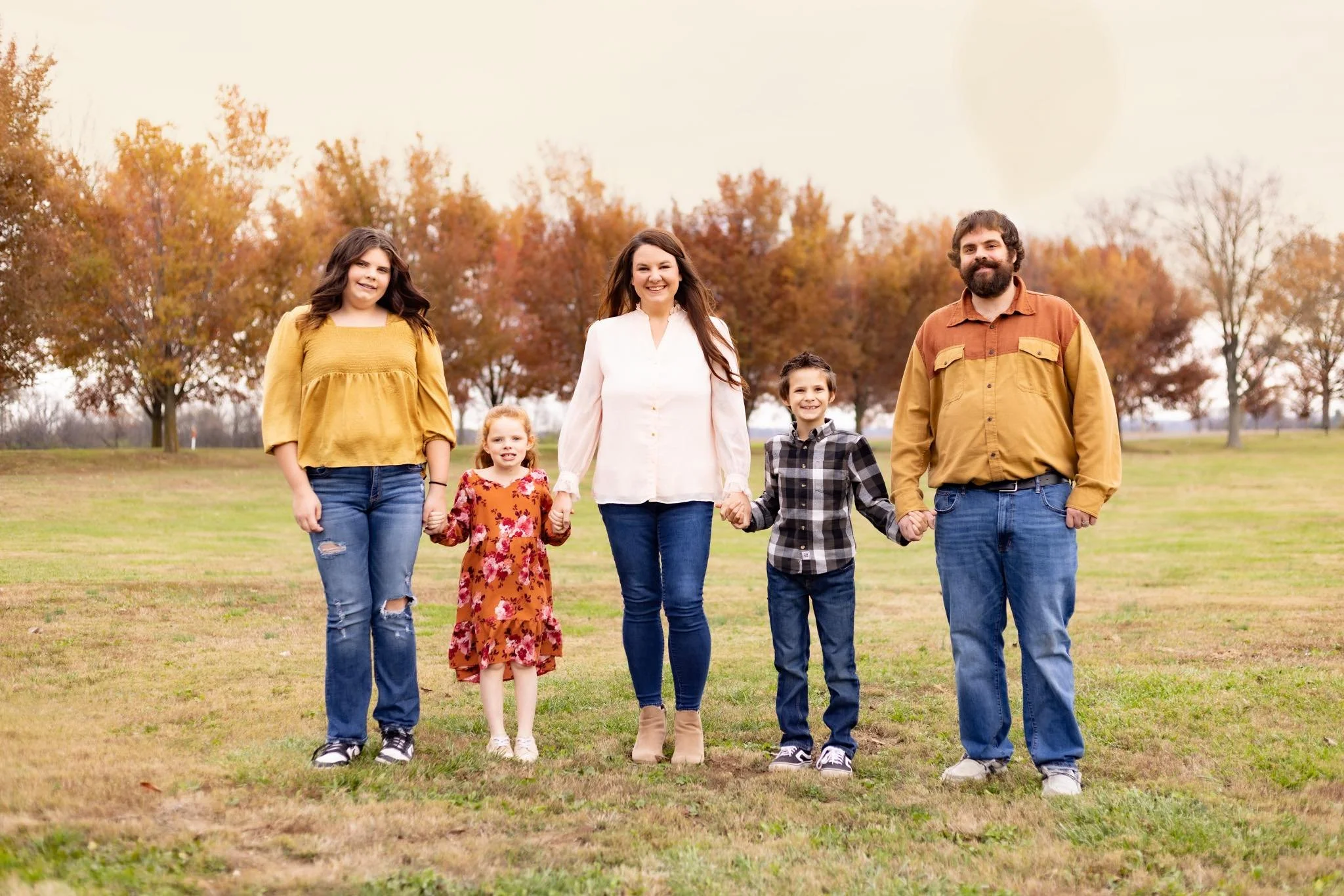 A family of five holding hands and walking in a park with autumn-colored trees in the background.