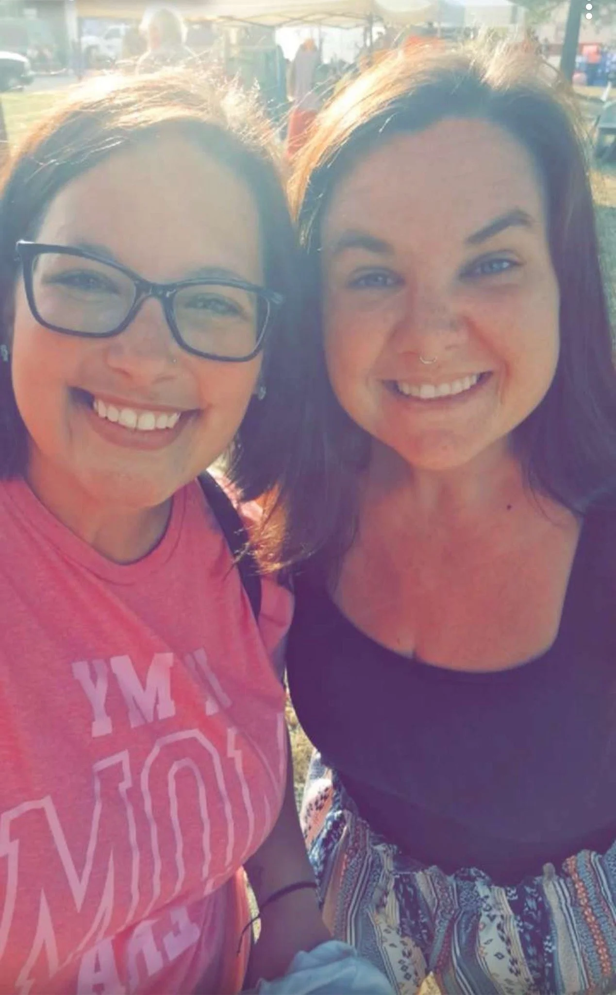 Two women smiling outdoors at a fair or festival, with tents and people in the background, sunlight shining on their faces.