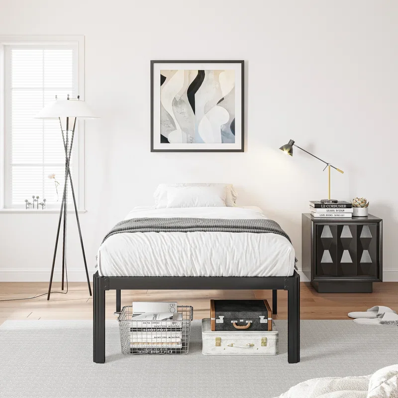 A minimalist bedroom with a black metal bed frame, white bedding, and a gray blanket. There's a abstract black and white artwork above the bed, a black bedside table with books and a gold desk lamp, a window with white blinds, a tripod floor lamp, wh