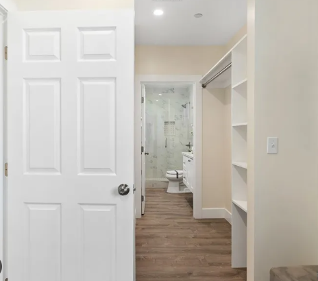 View of a bathroom from a room with an open closet door, showing a shower, toilet, and vanity in the background.