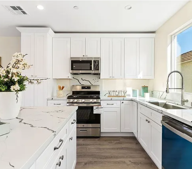 Modern white kitchen with marble countertops, stainless steel appliances, and a large window with a view outside.