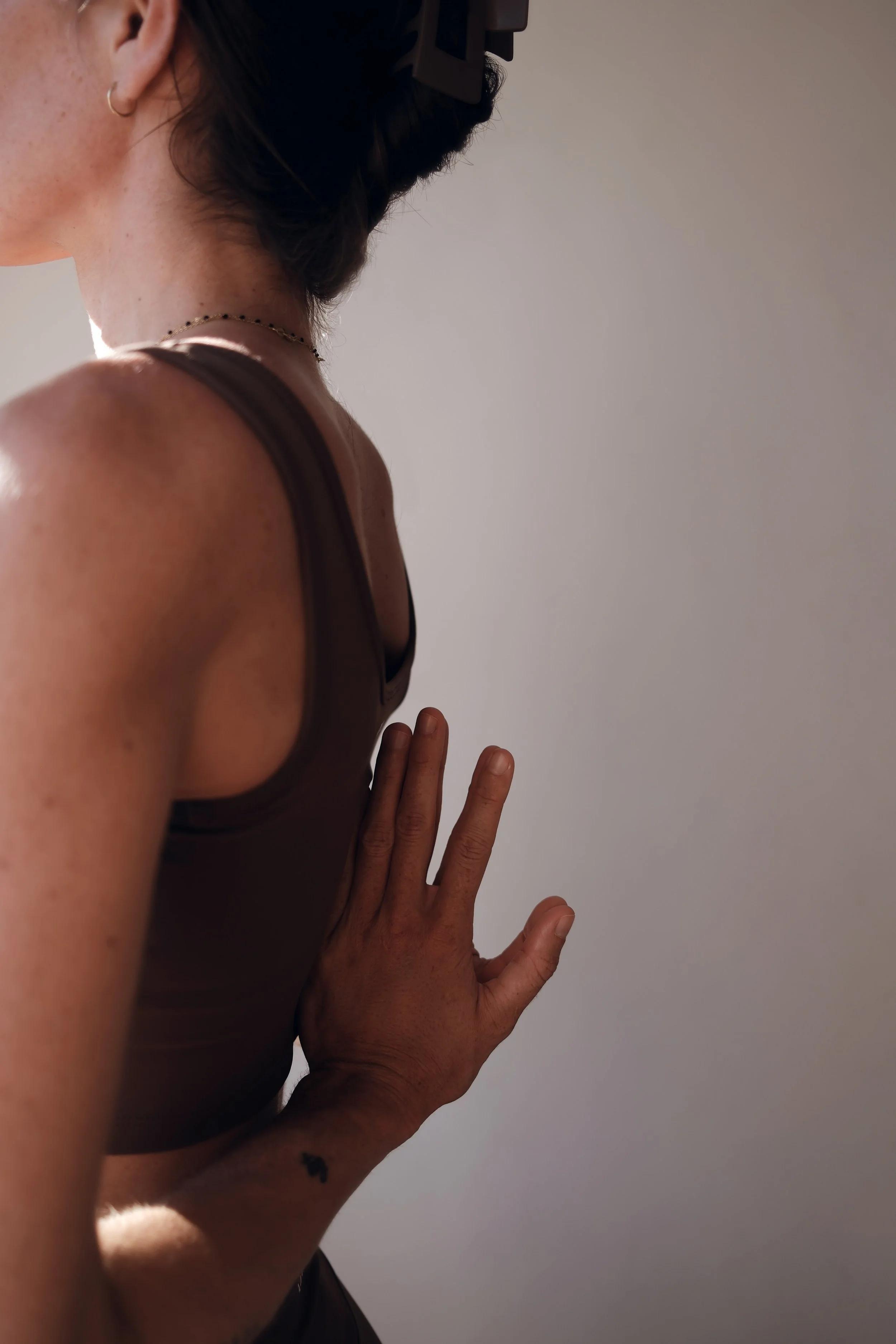 A person with short hair wearing a brown tank top, a necklace, and a small earring, pressing their hand flat against a plain wall.