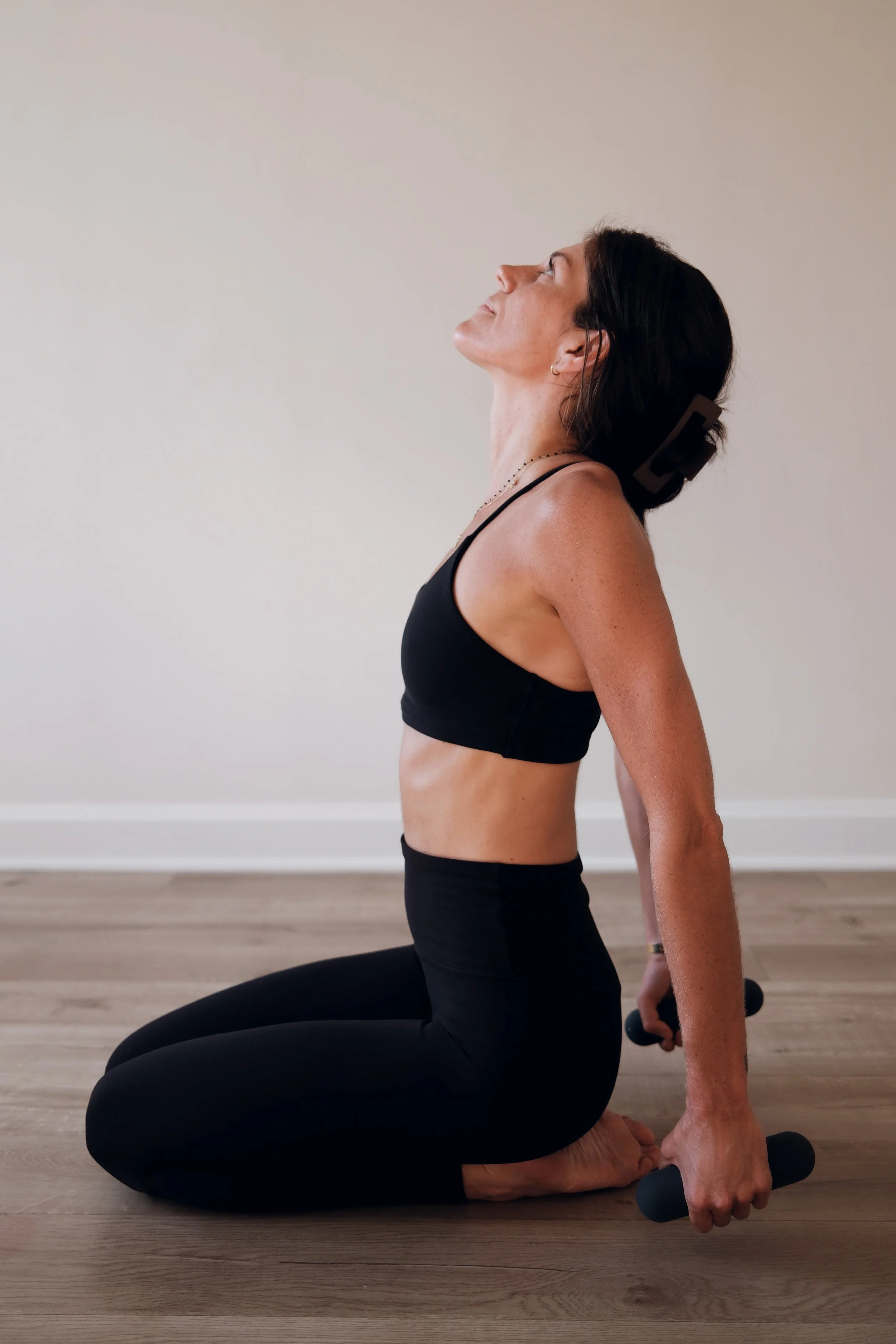 Woman in black workout attire doing yoga or stretching on the floor, holding dumbbells behind her back, with her head tilted back and eyes closed.