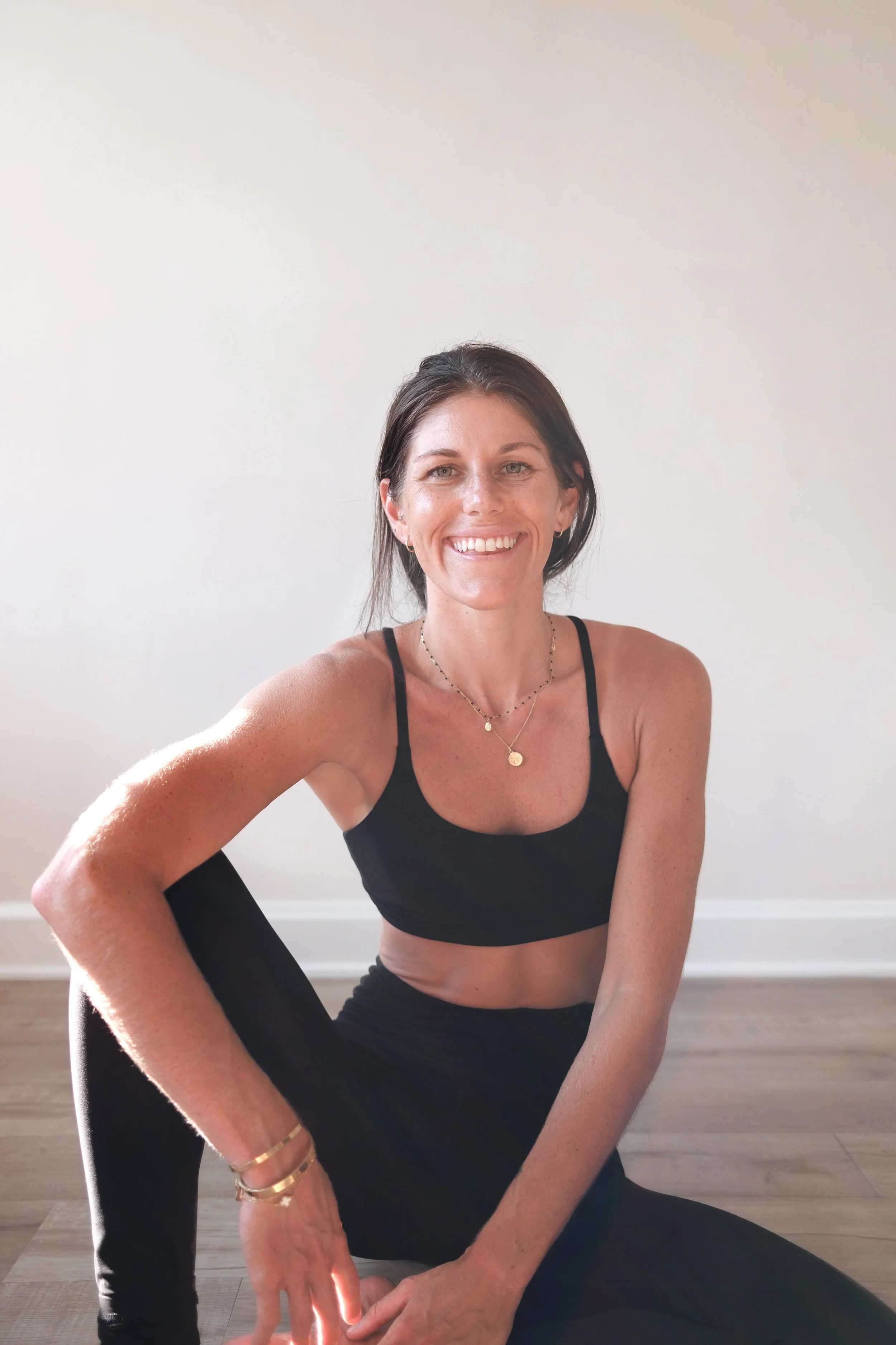 A woman smiling while sitting on the floor in a room with hardwood flooring and white walls.