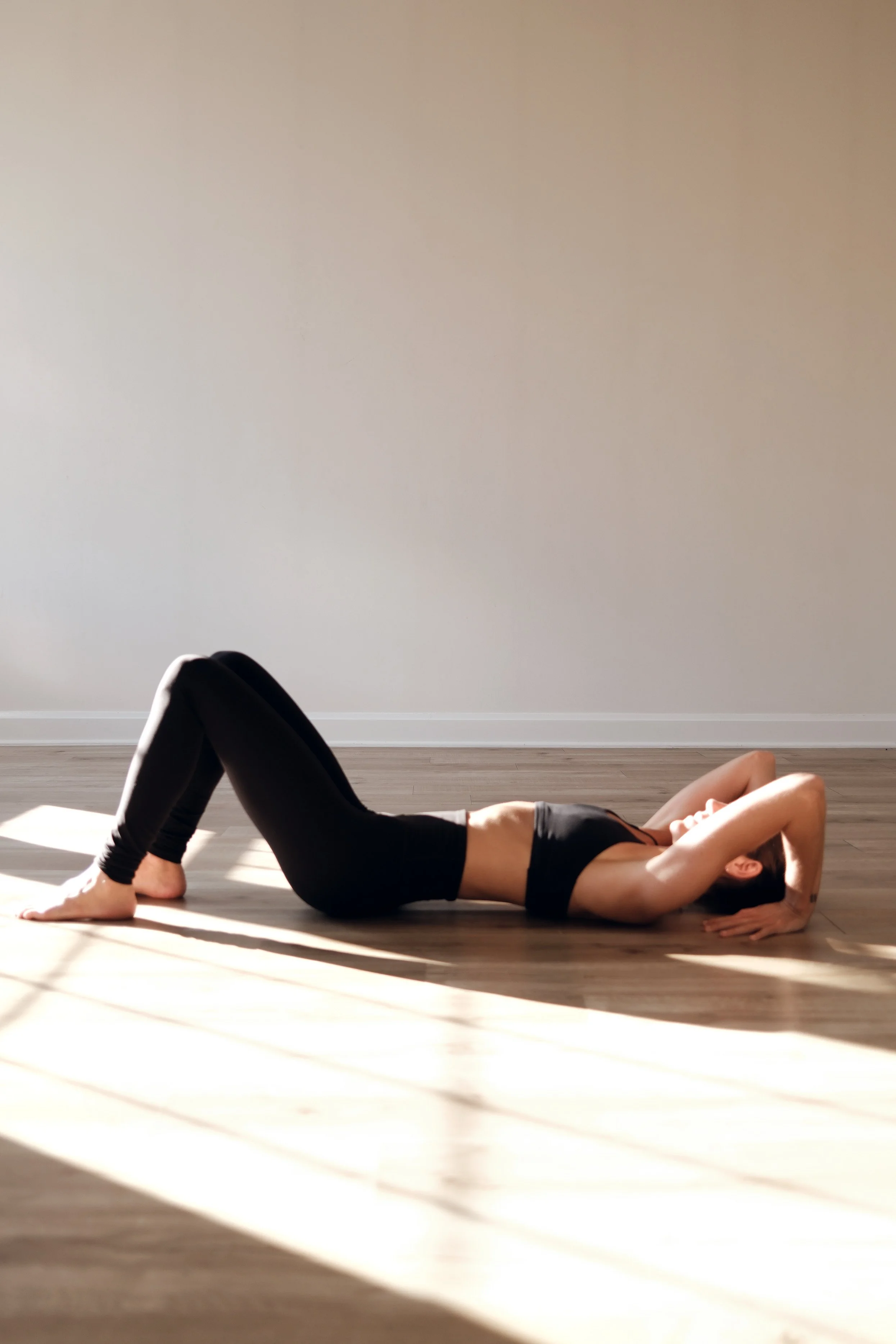 A woman practicing yoga indoors on a wooden floor, lying on her back with knees bent and hands behind her head, in a relaxed pose.