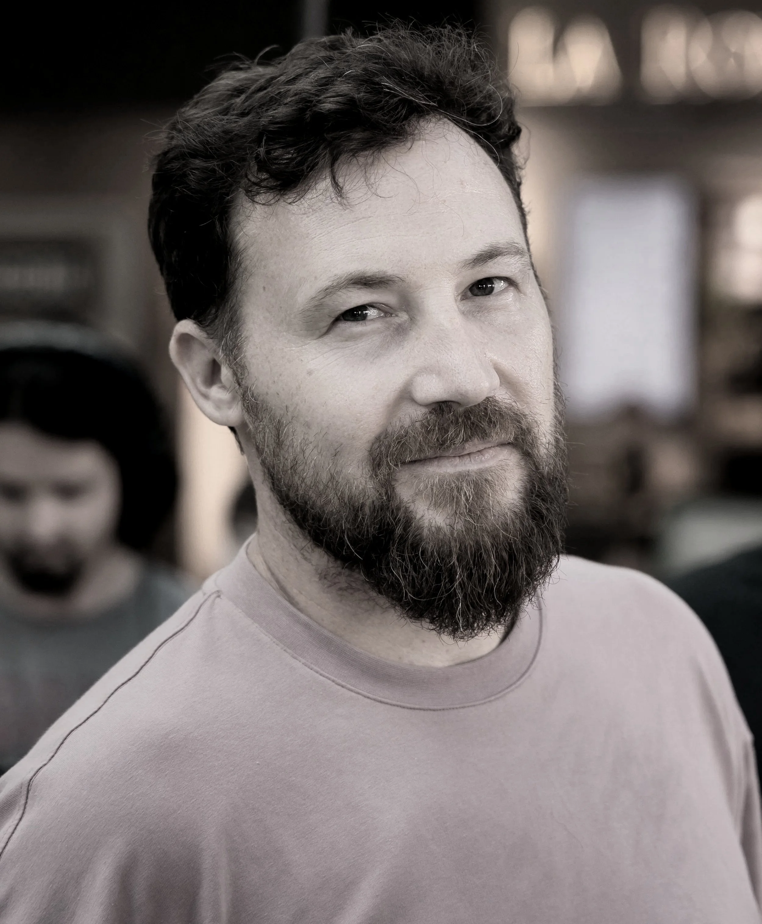 A black and white photo of a man with a beard and mustache, wearing a light-colored t-shirt, smiling slightly, with a out-of-focus person in the background.