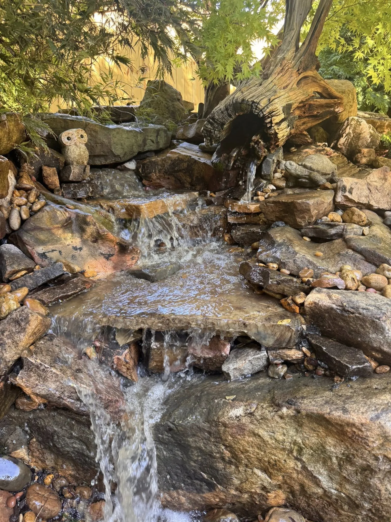 A small garden stream flowing over rocks, with a rock shaped like an owl and a weathered tree branch waterfall feature, surrounded by green foliage and pebbles.