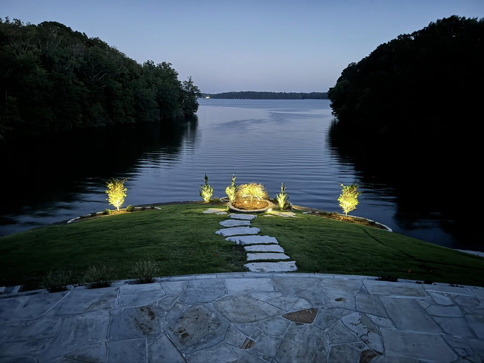 Night view of a lake with trees on both sides and a small grassy area with a pathway and garden lights illuminating shrubs and trees in the foreground.