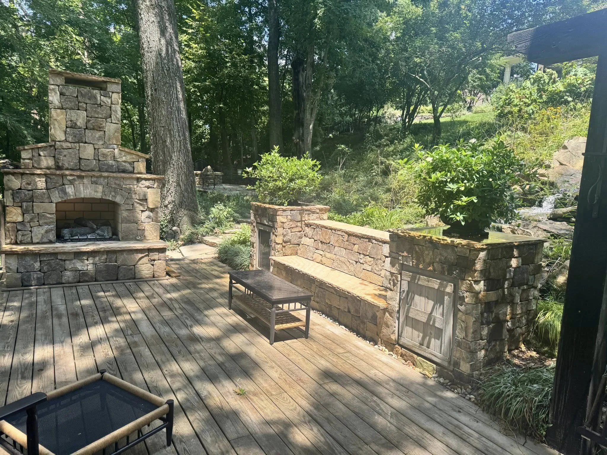 A wooden deck in a backyard with a stone fireplace, planters, and lush green trees and plants in the background.