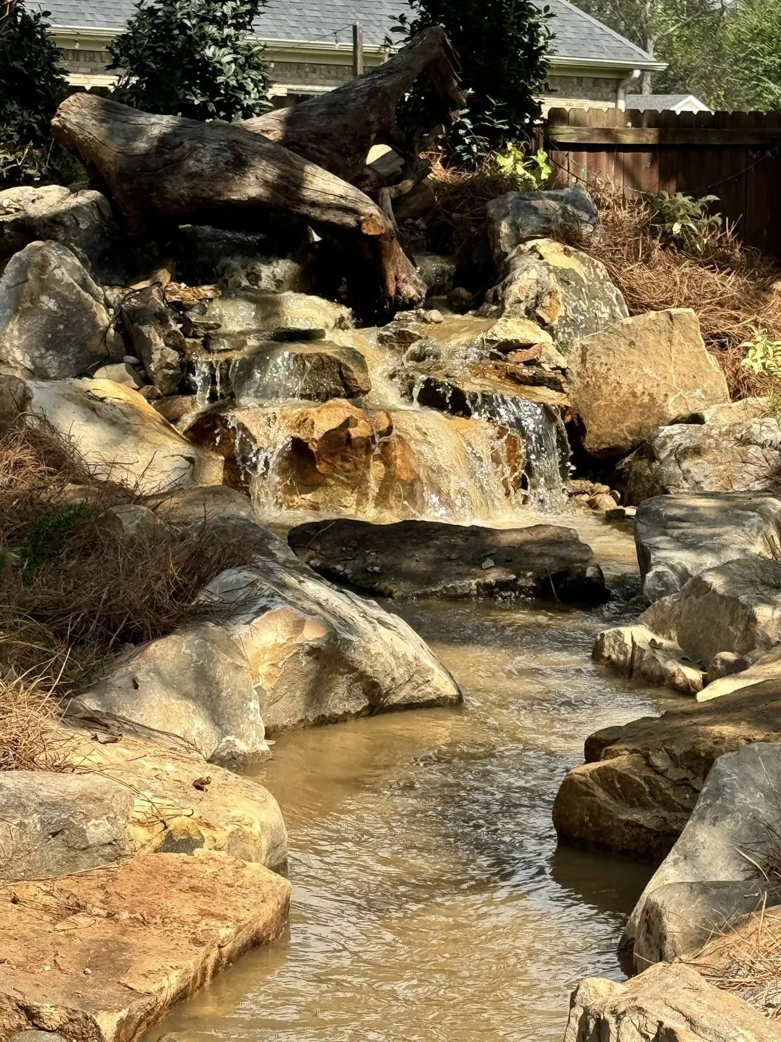 A small man-made waterfall cascading over rocks and surrounded by trees and shrubs in a backyard.