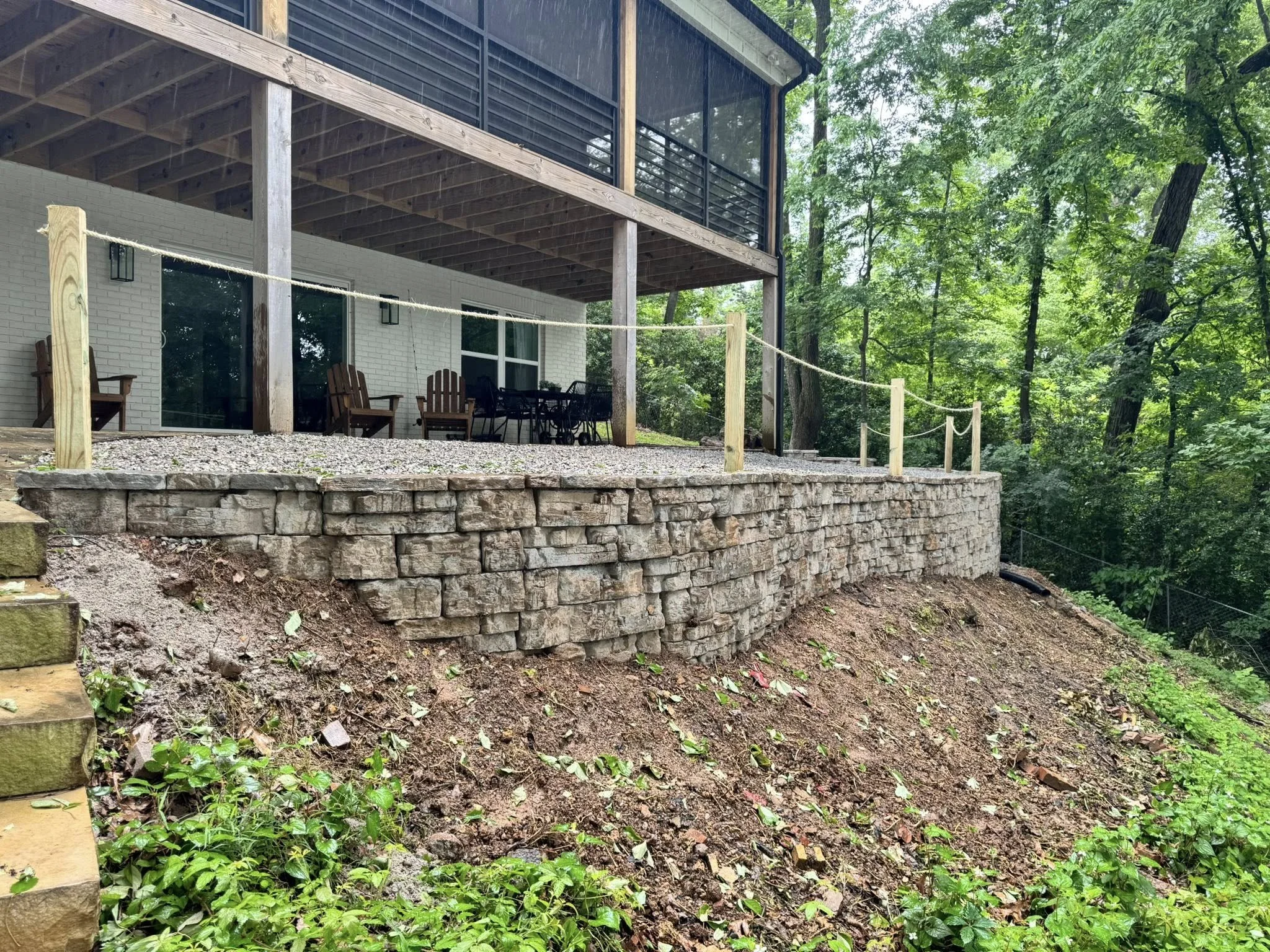 A backyard patio with a stone retaining wall and a safety rope fence on wooden posts. The patio is attached to a modern house with large glass sliding doors and a balcony above. There are wooden chairs and a table on the patio, and the area is surrou