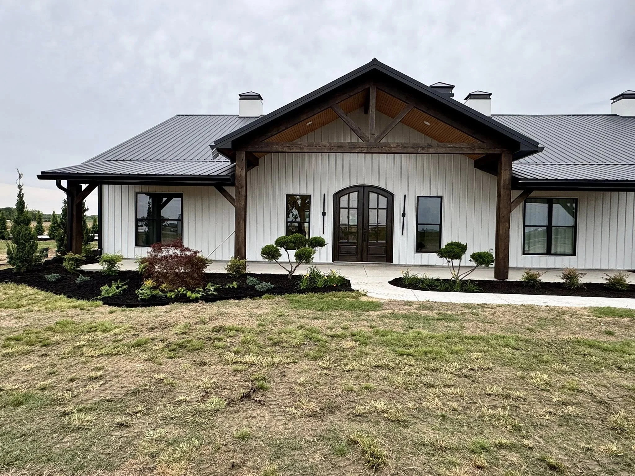 A modern farmhouse with a metal roof, white vertical siding, and a central arched door. The house has dark trim and small landscaped garden beds with shrubs and plants in front.