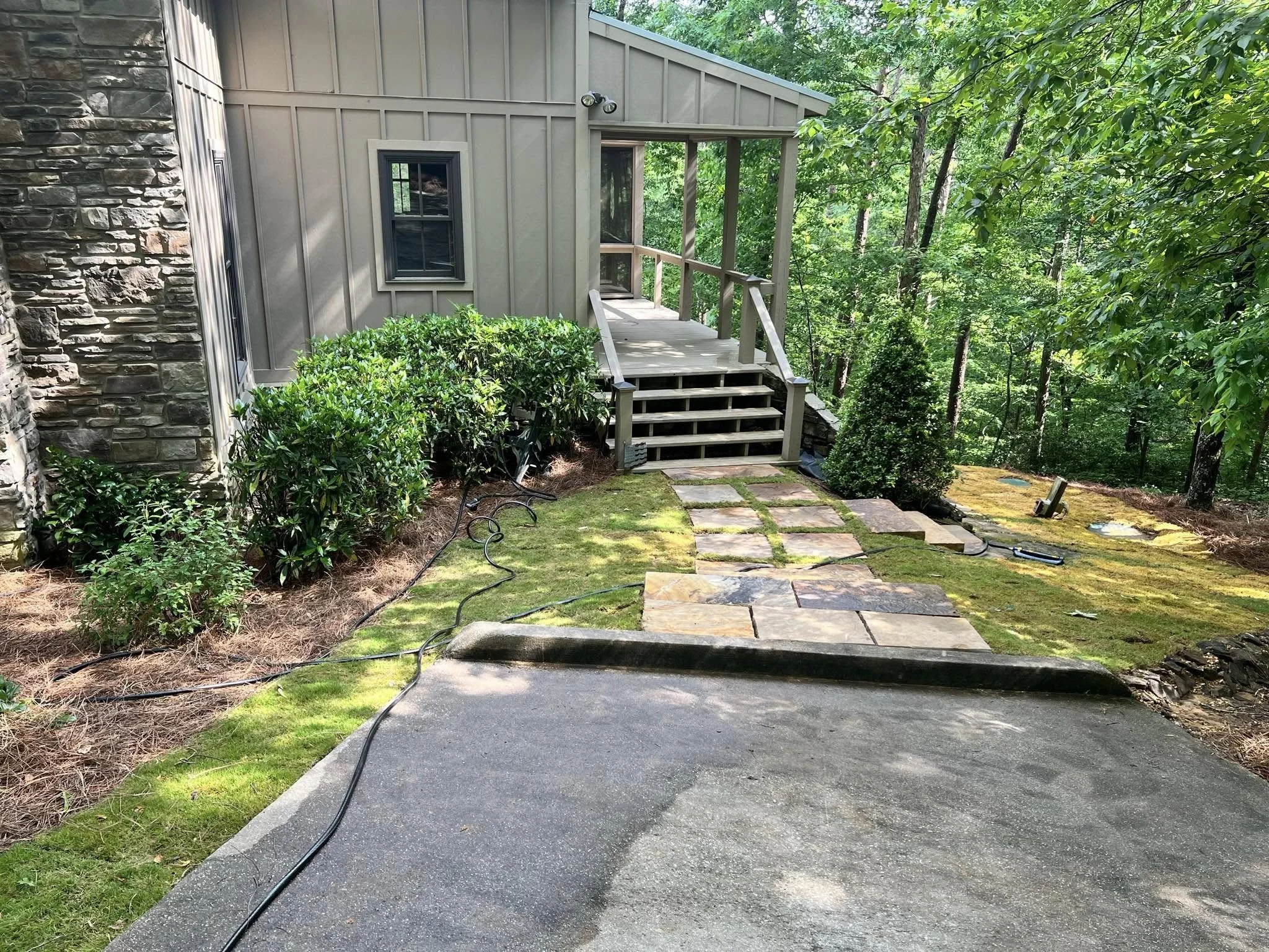 Stone pathway leading to stairs up to a house with a wooden porch, surrounded by green bushes and trees in a wooded yard.