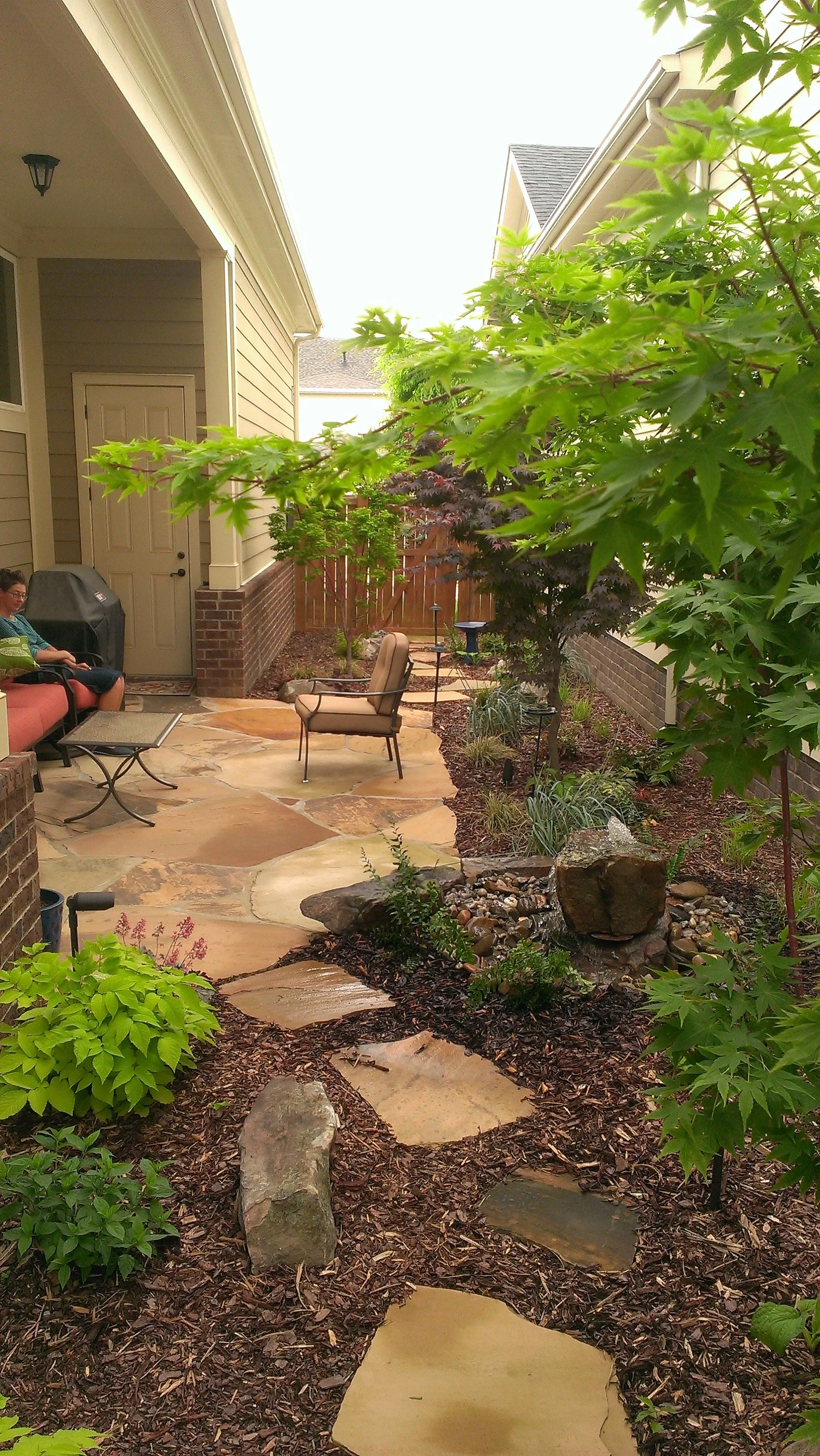 A backyard patio with a stone pathway, two chairs, a table, a tree, and some shrubs, with part of a house and a wooden fence visible.