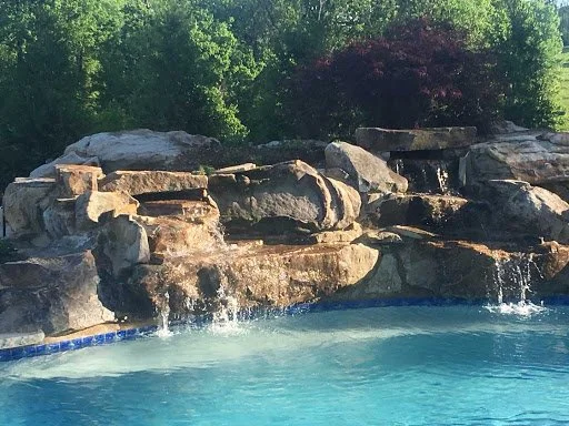 A backyard pool surrounded by rocks and lush green trees.