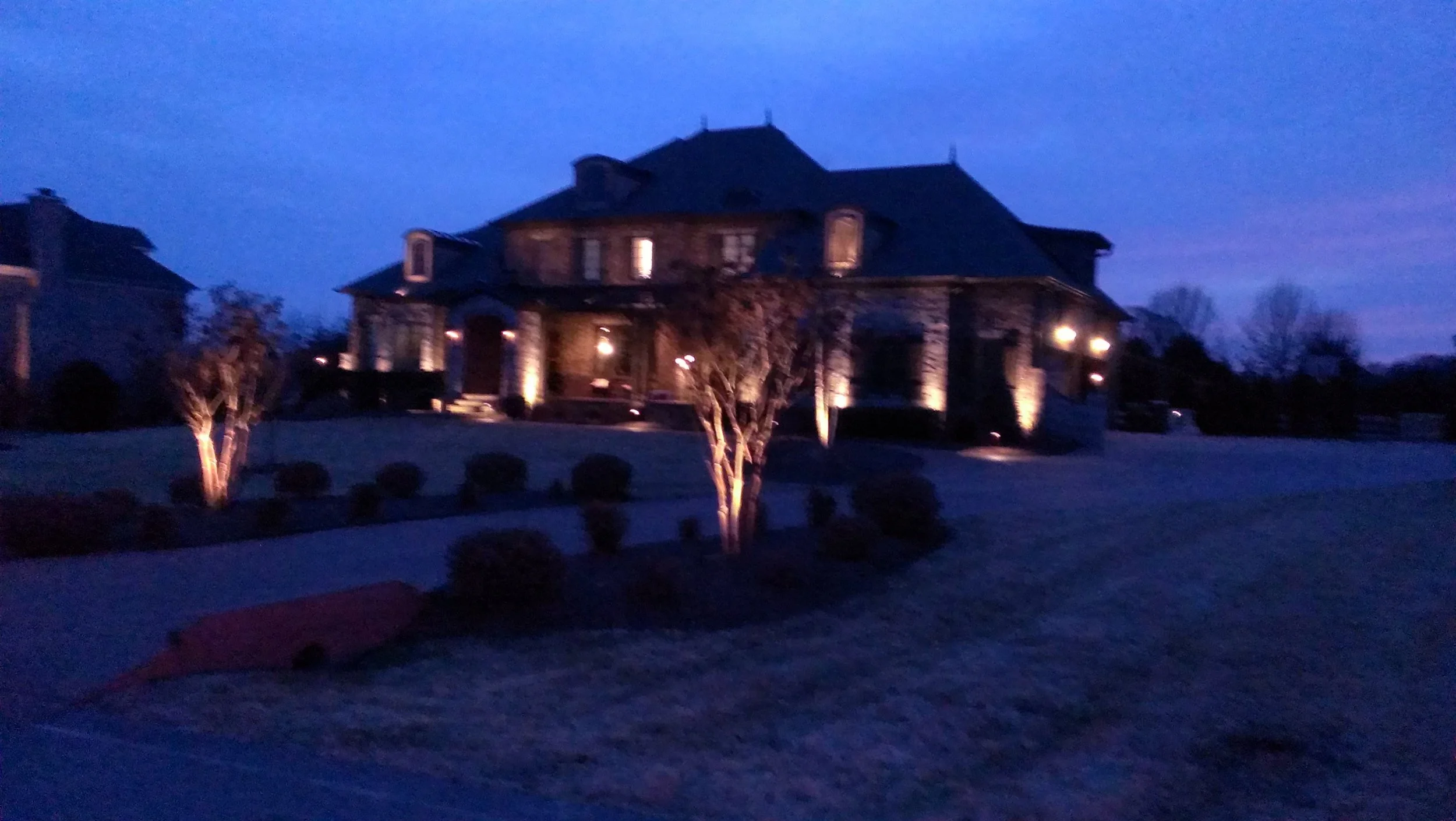 Large brick house illuminated at night with exterior lights, trees in front, and a driveway on the left.
