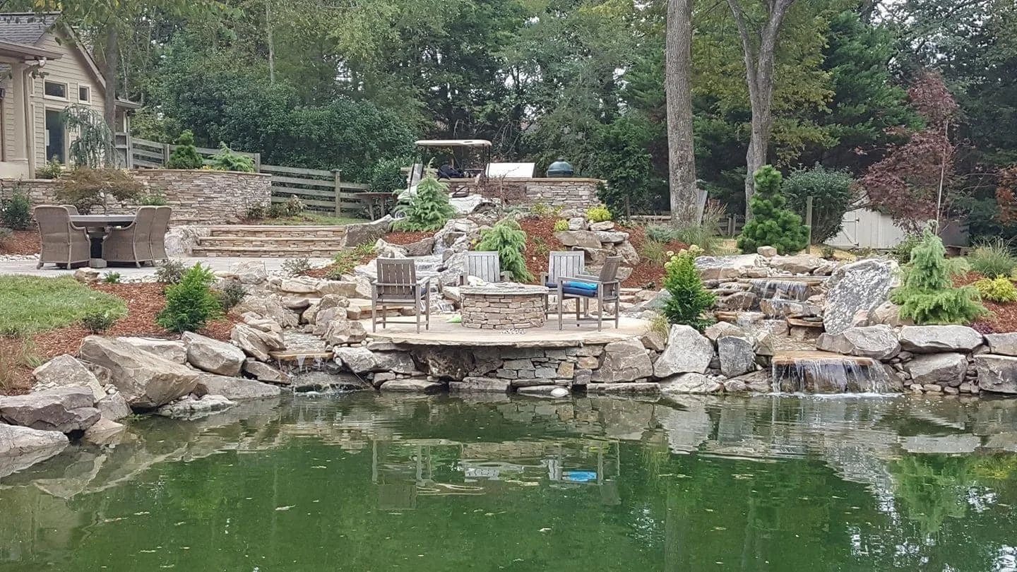 A backyard with a pond, waterfalls, rocks, trees, potted plants, and outdoor furniture including chairs and a table.