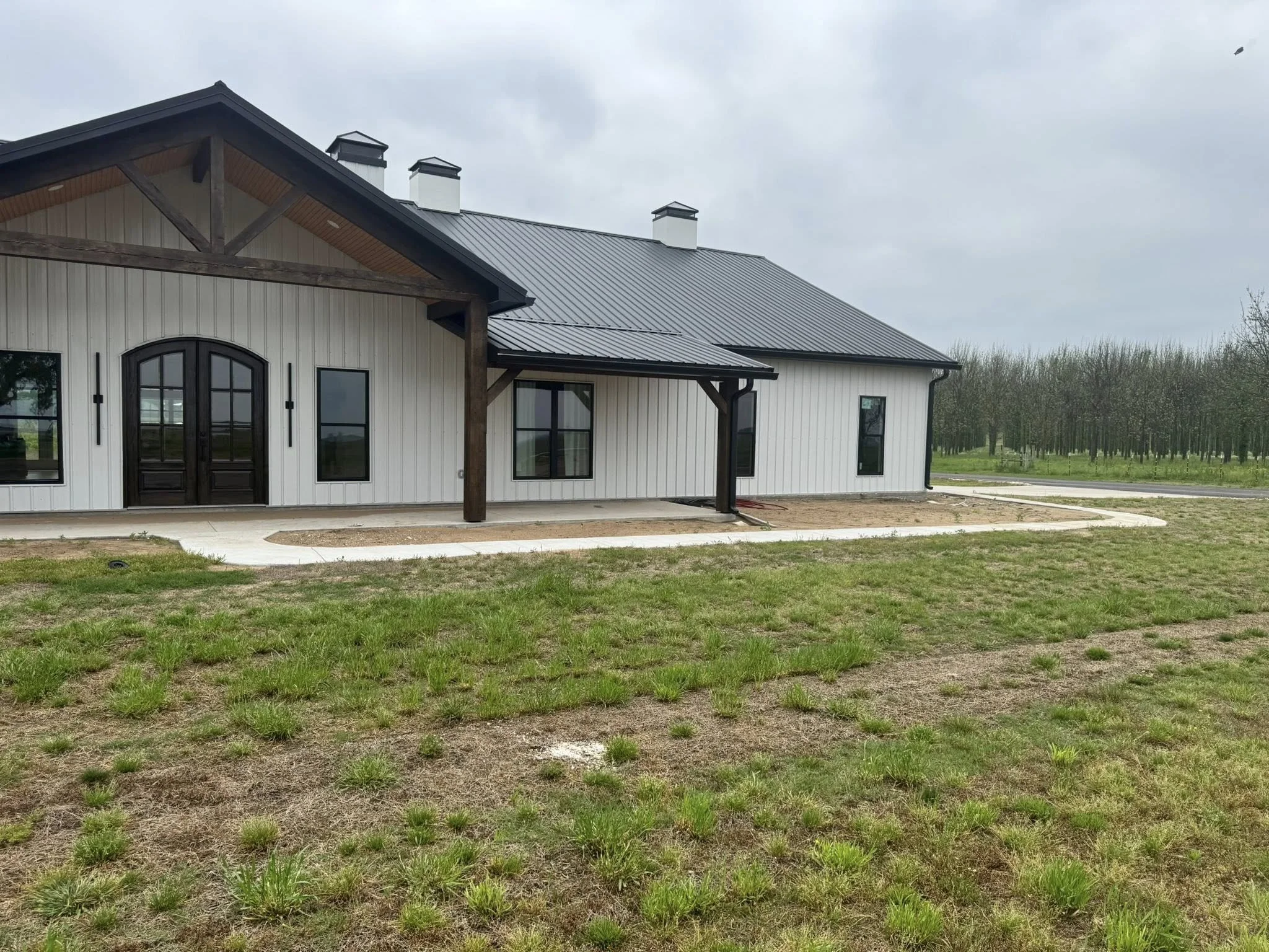 A modern farmhouse-style building with white vertical siding, black door, black-framed windows, a black metal roof, and a small covered porch area supported by wooden beams. The yard has a concrete pathway and patches of grass, with an overcast sky a