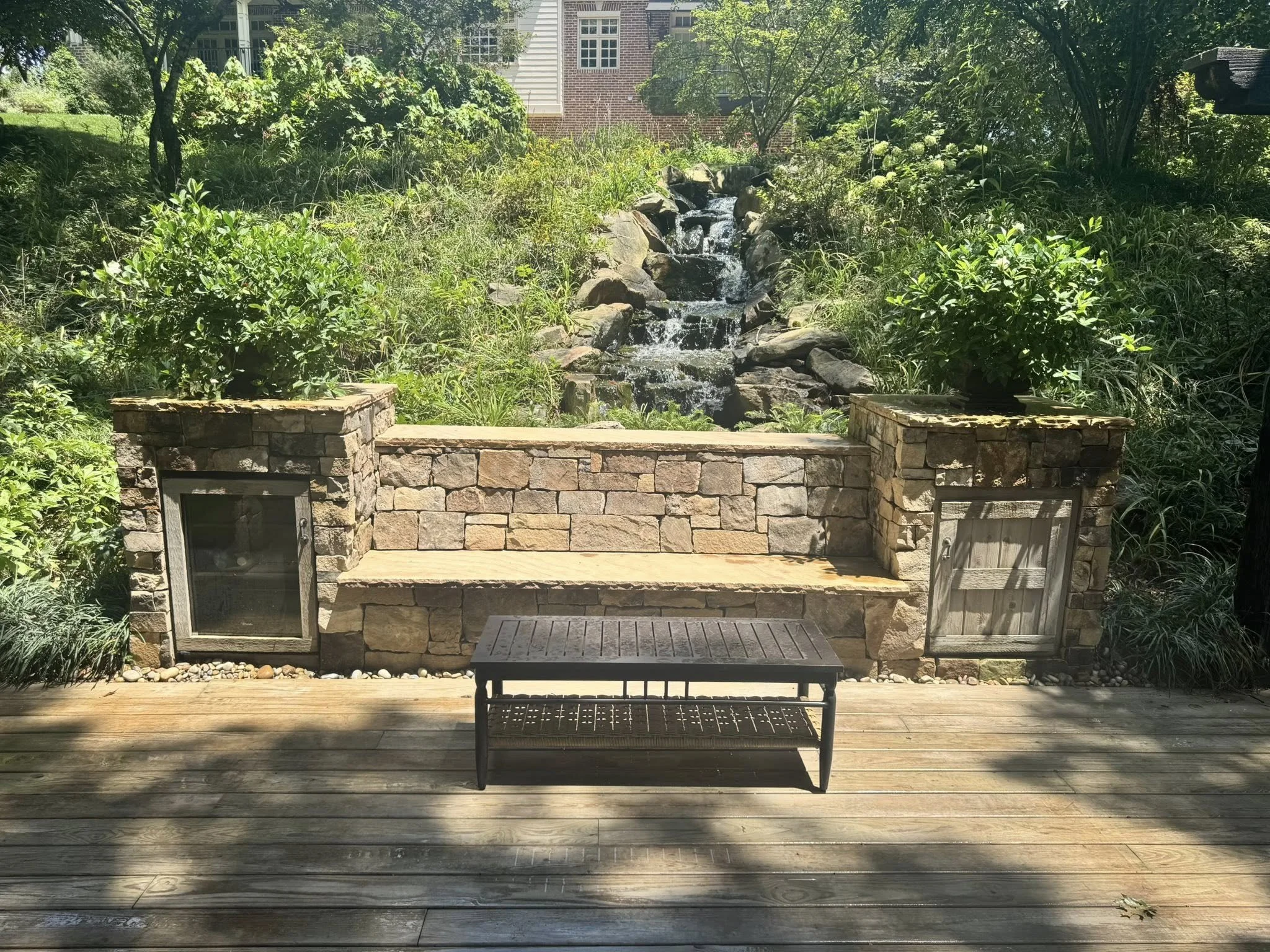 Stone and brick outdoor water feature with waterfall, flanked by shrubs, on a wooden deck with a metal bench in front.