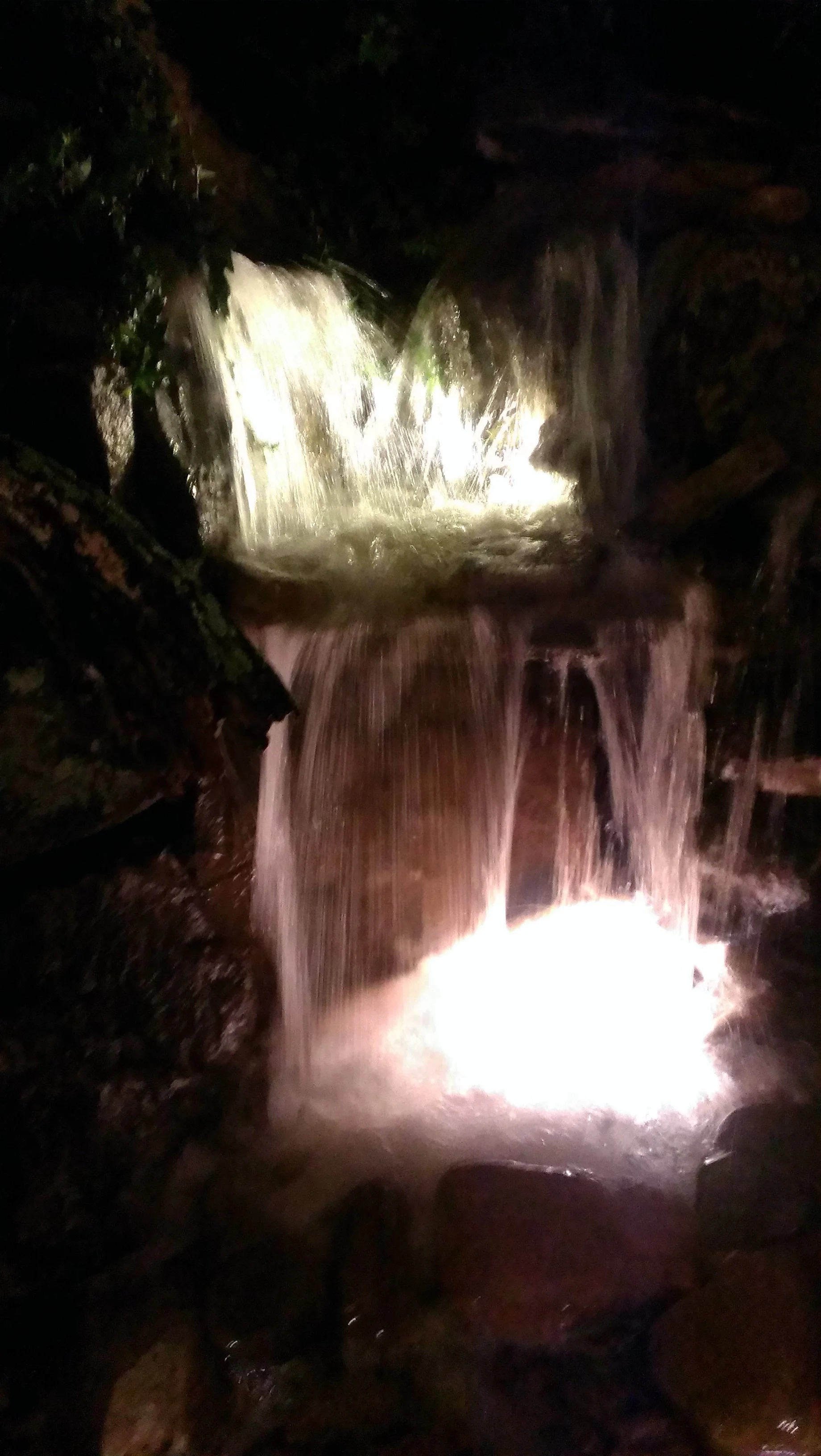 Nighttime view of a small waterfall with illuminated flowing water over rocks and surrounded by dark rocks and vegetation.