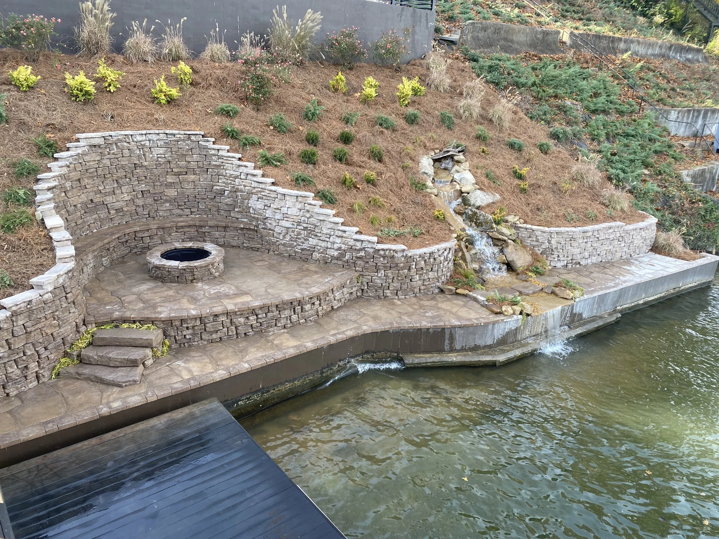 Outdoor backyard area with a stone patio, built-in fire pit, and a waterfall feature flowing into a pond, surrounded by landscaped rock and plants.