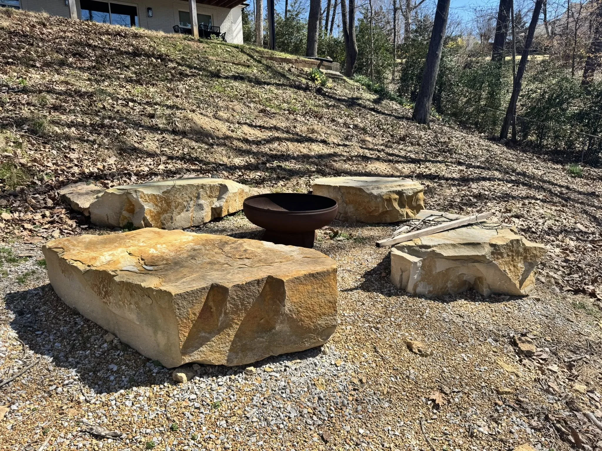 Large beige and light brown rocks arranged in a semi-circle outdoors with a fire pit in the center, on a gravel surface, with trees and a house in the background.