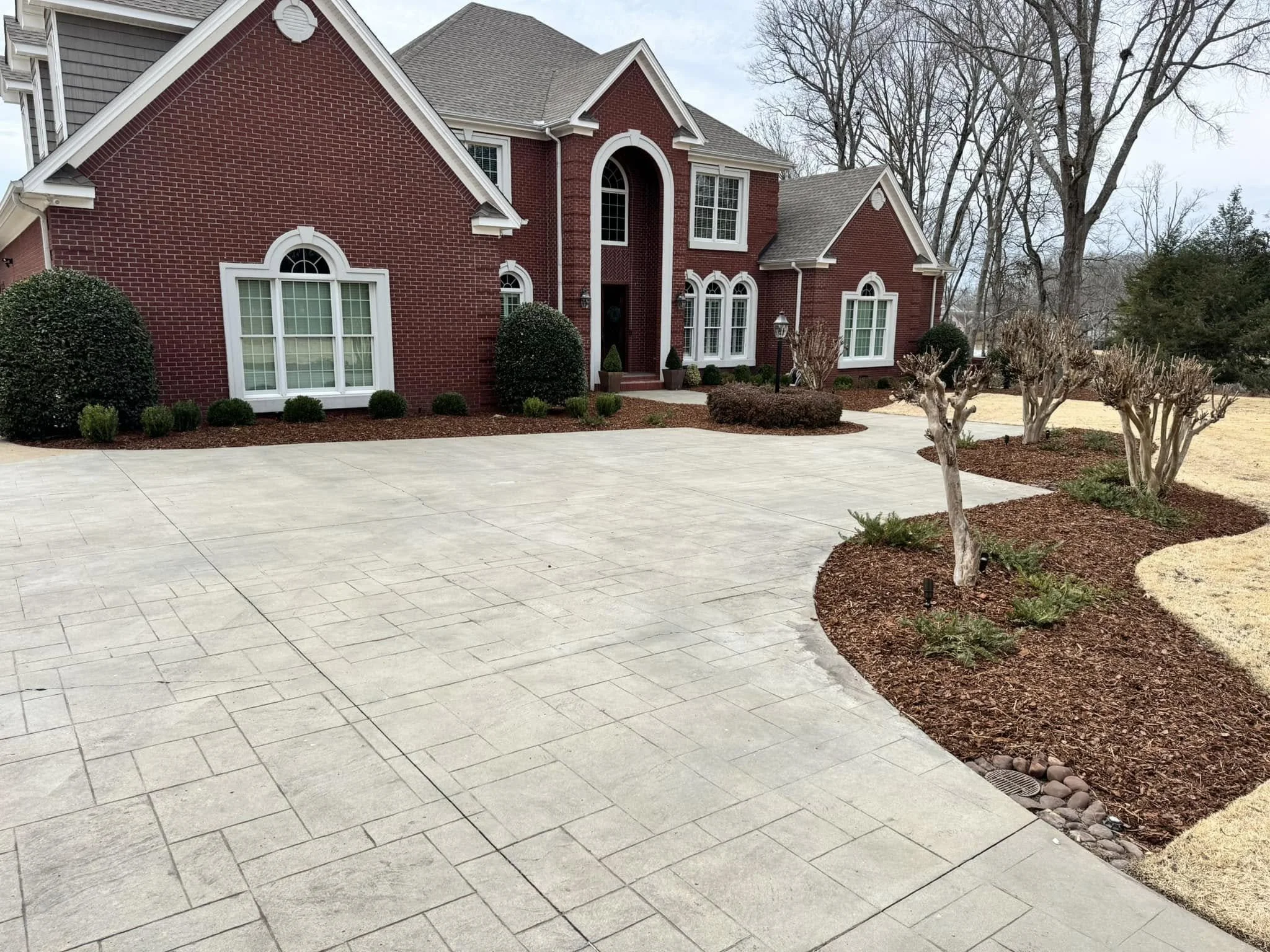 Front yard of a red brick house with a wide concrete driveway, manicured bushes, leafless trees, and a lawn with brown grass.