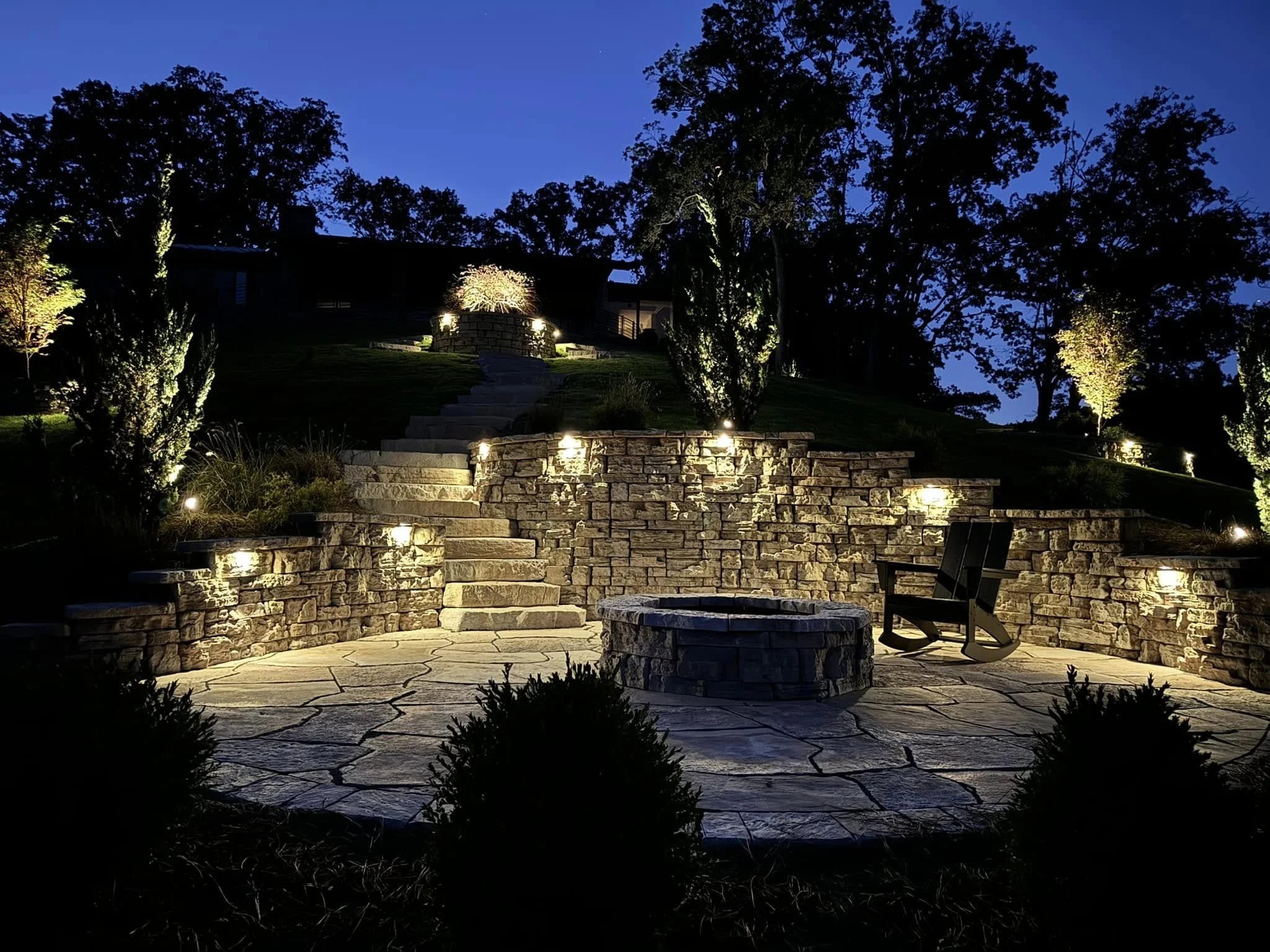 Nighttime view of a lit stone patio with a fire pit, surrounded by a stone wall, illuminated pathway, and trees in the background.