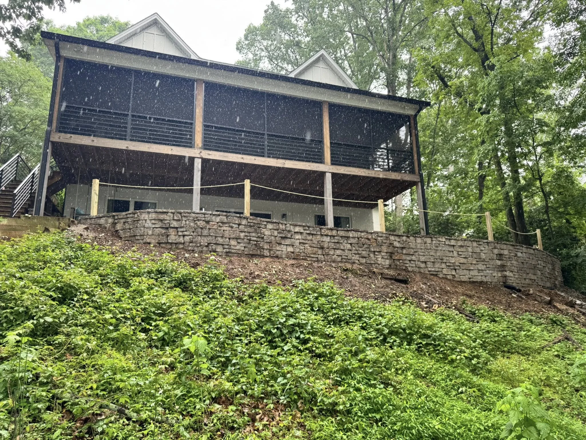 A house on a hill with a screened-in porch, stone retaining wall, and lush green trees surrounding it during rain.