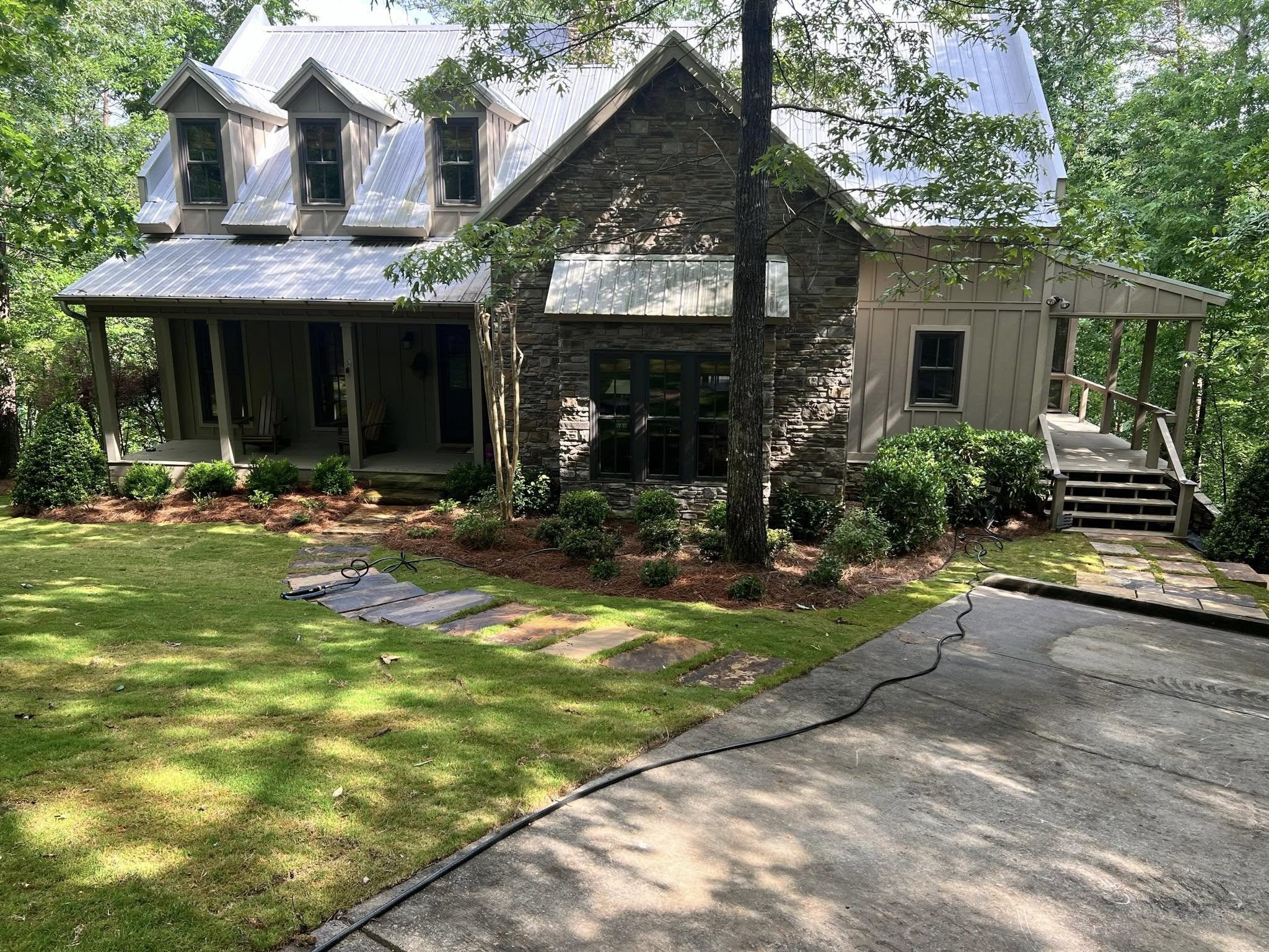 A house with a slate roof and a combination of stone and siding exterior, surrounded by trees and a well-maintained lawn. There is a porch with patio chairs and a set of stairs leading to a side entrance. A concrete driveway with a hose running acros