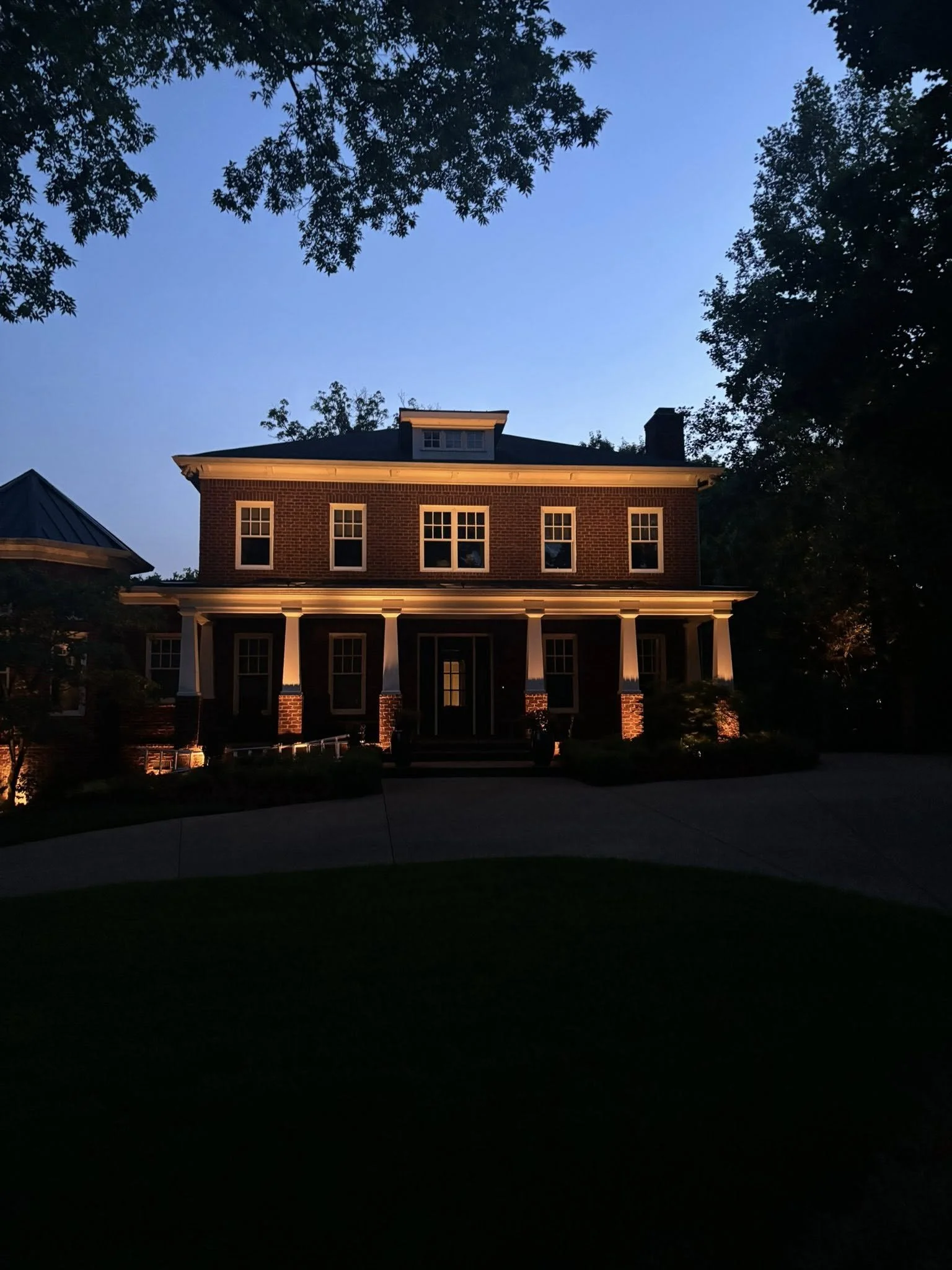 A large brick house at dusk with exterior lighting illuminating the front porch and facade, surrounded by trees.